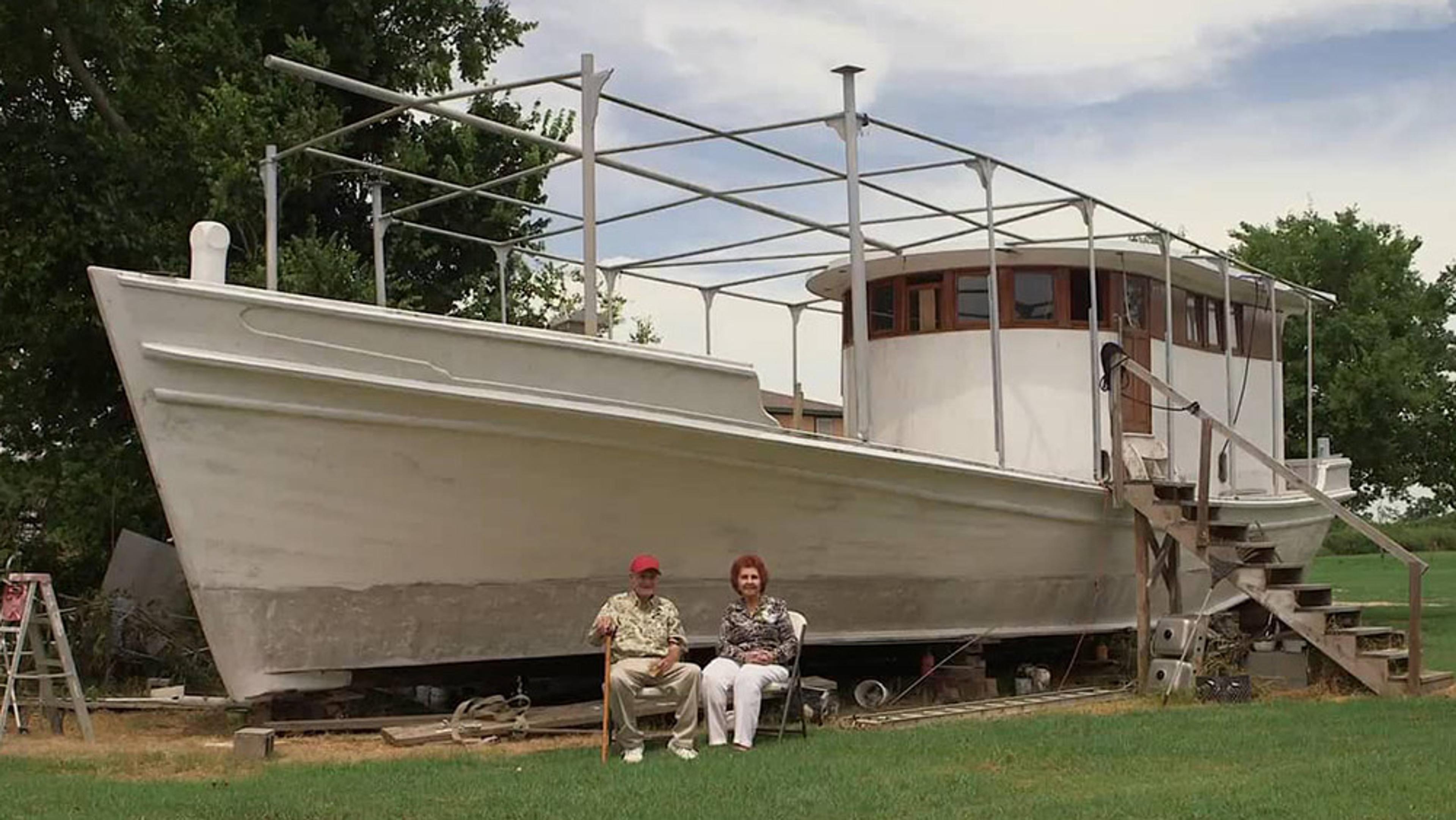 A large white boat on land with an elderly man and woman sitting in chairs on grass in front of it.