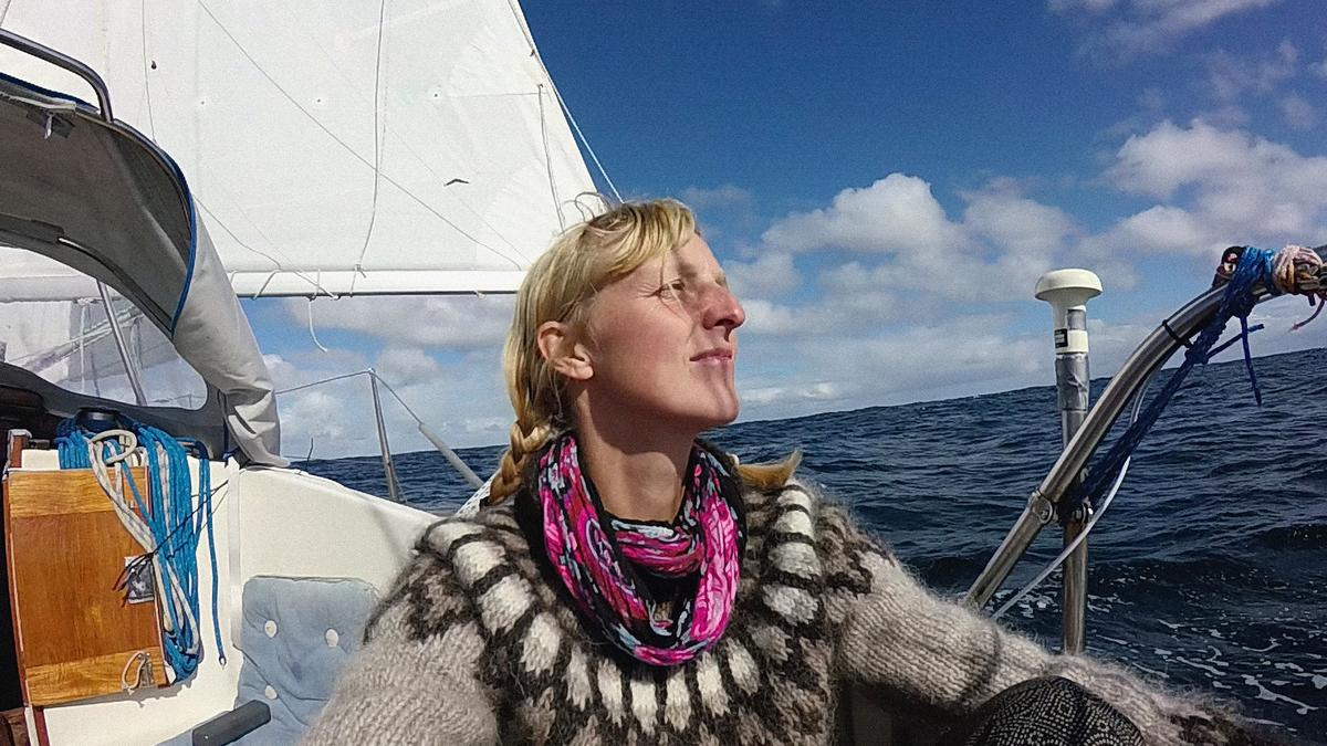 Photo of a woman with braided hair on a sailboat in open sea, wearing a patterned jumper and colourful scarf, enjoying the sun.