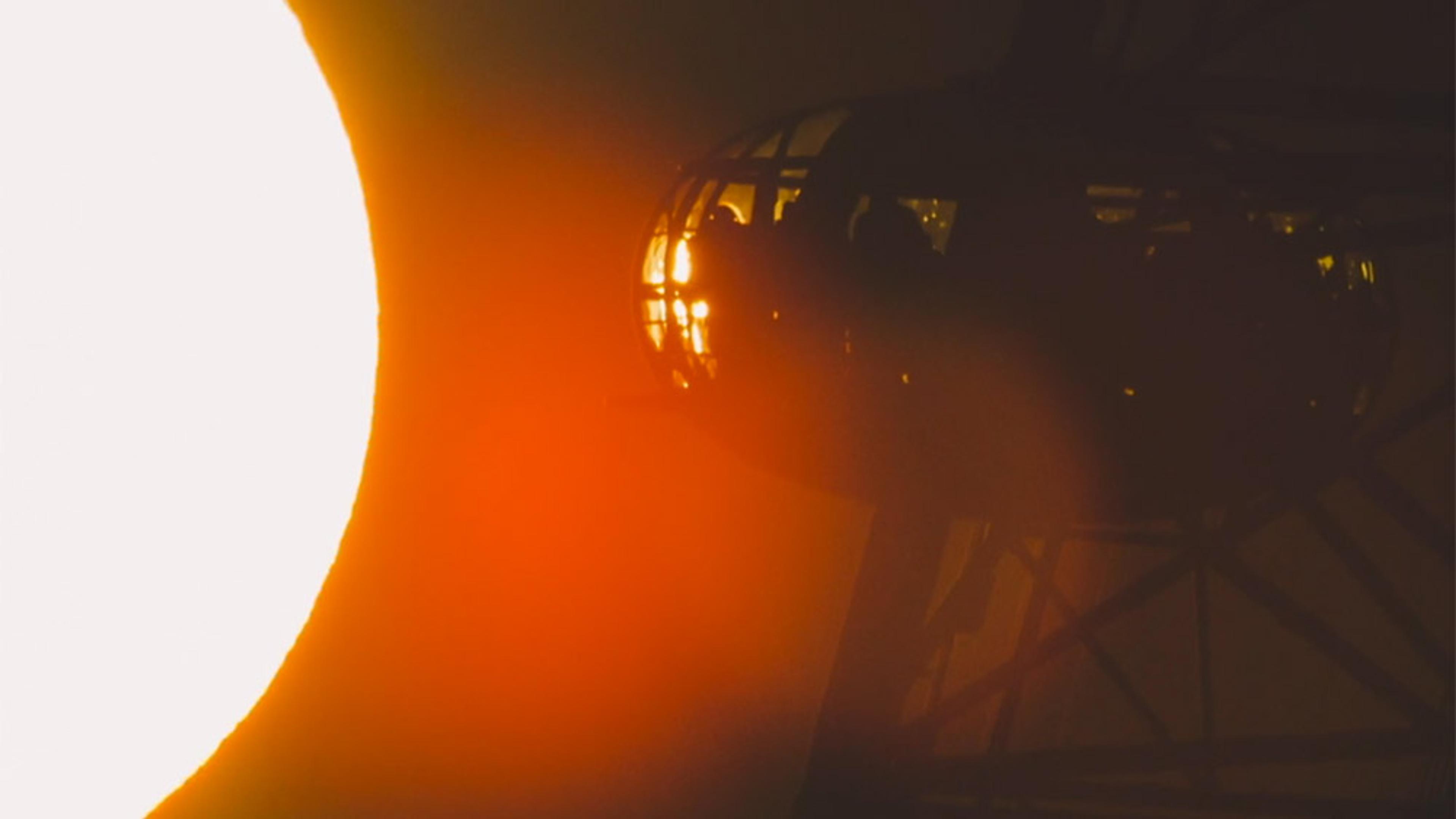 A bright sun partially obscuring a ferris wheel cabin with silhouetted passengers against an orange-yellow gradient sky.