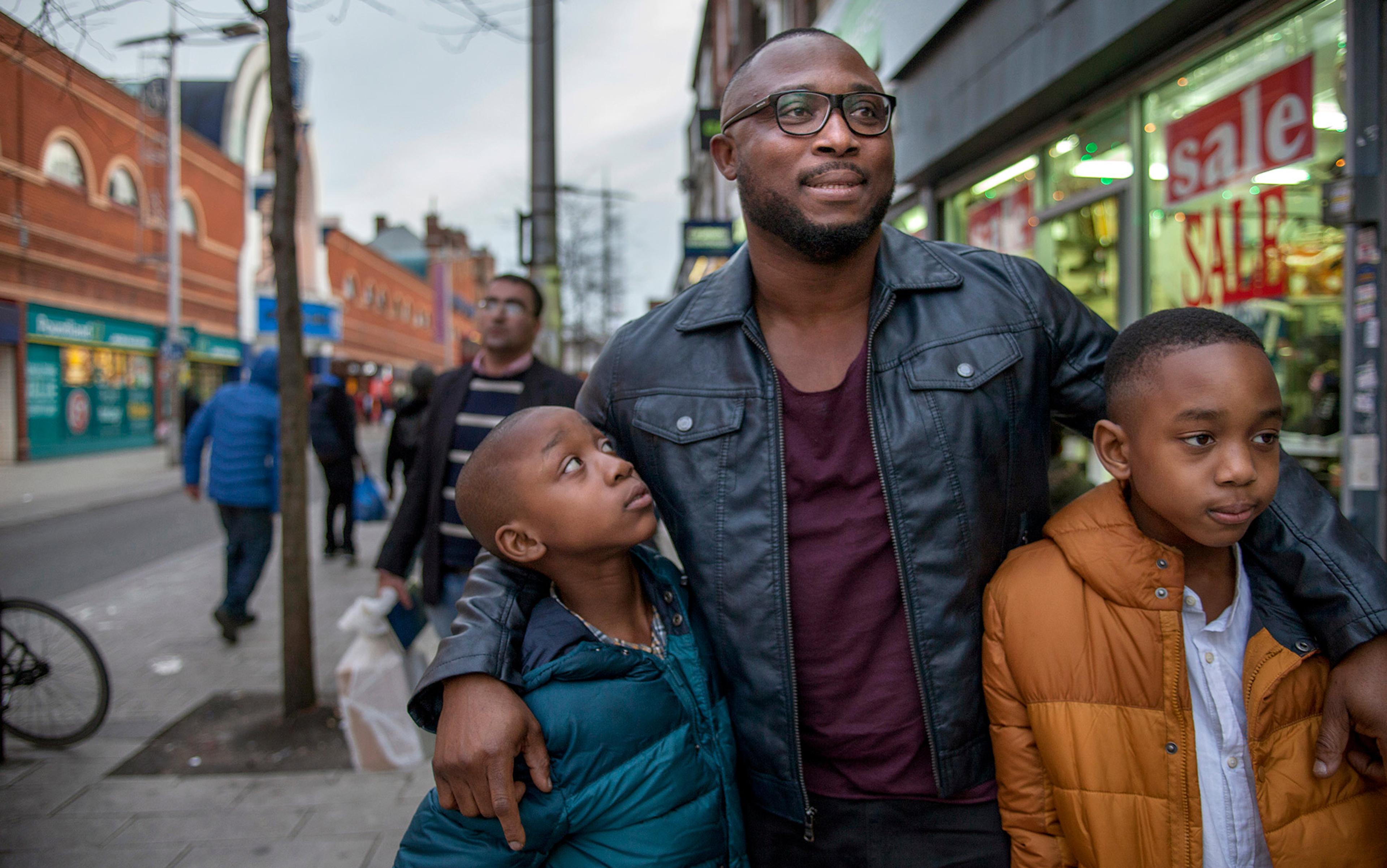 A man with glasses and two boys walk together on a city street along a row of shops.
