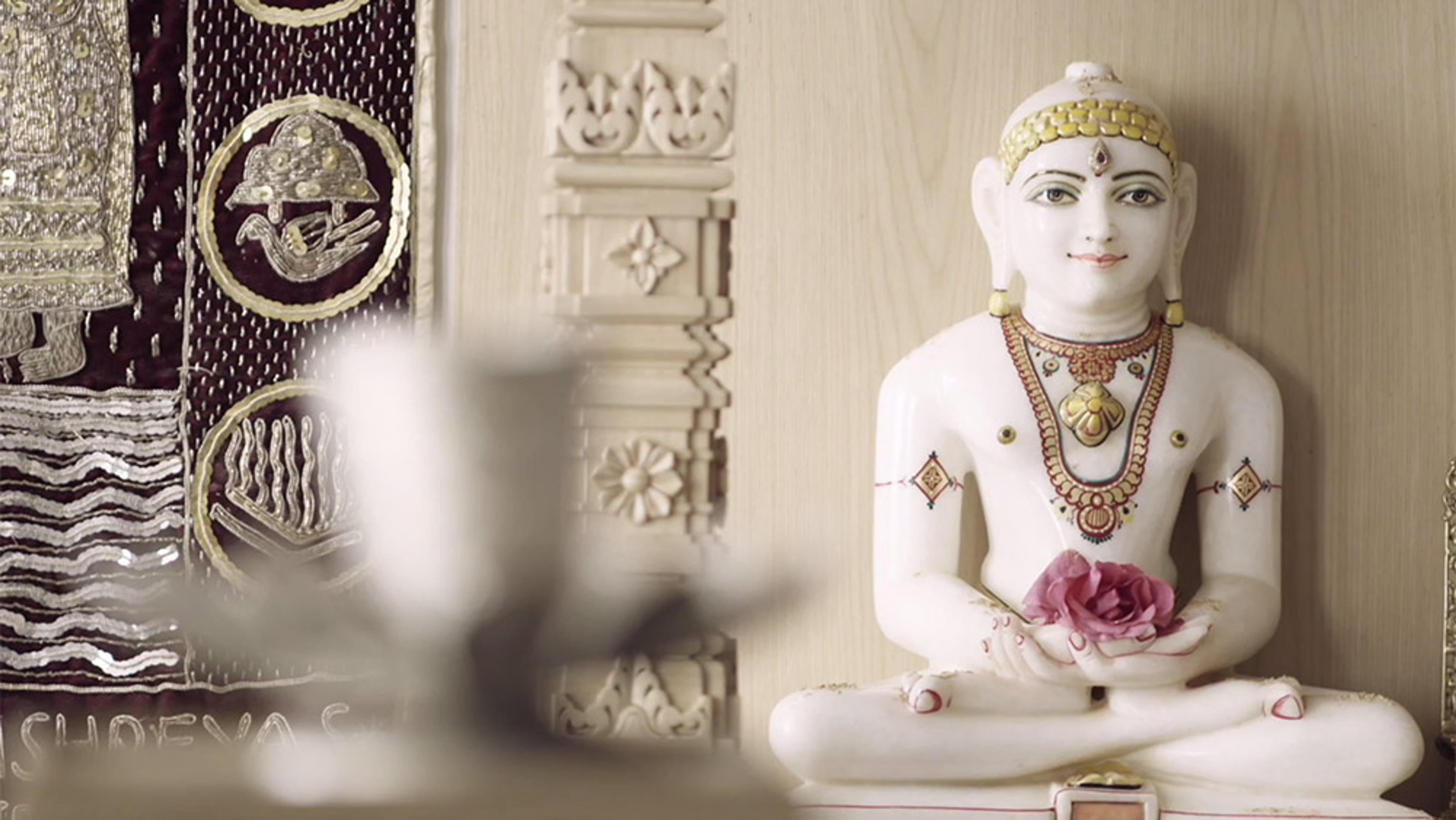 A white statue of a cross-legged figure holding a pink lotus, with intricate wall carvings and a tapestry in the background.