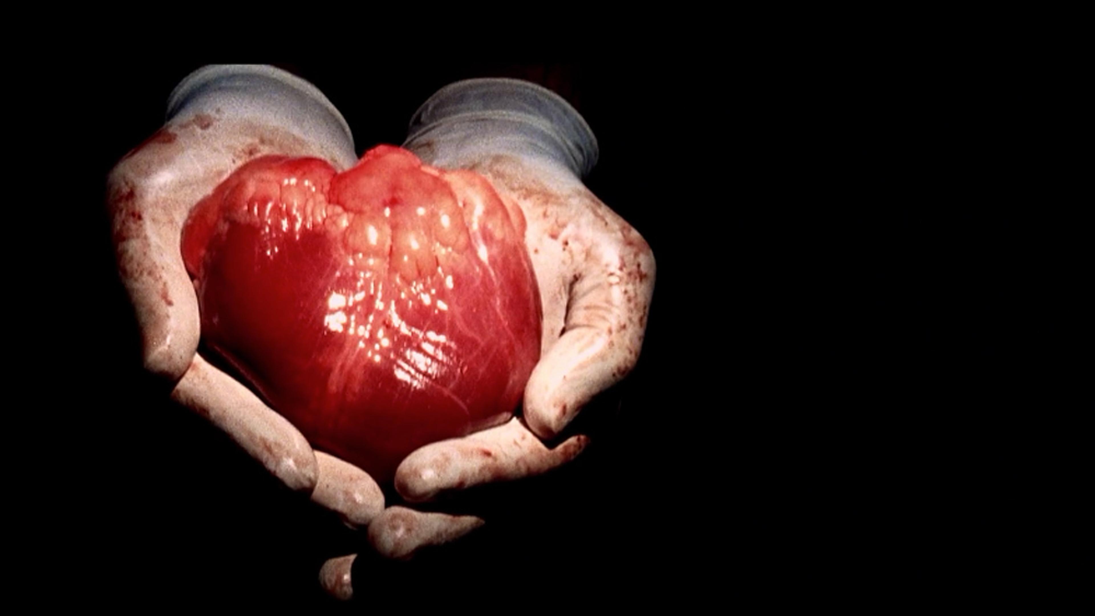 Gloved hands holding a human heart against a dark background. The heart is freshly extracted, with visible bloodstains on the gloves.