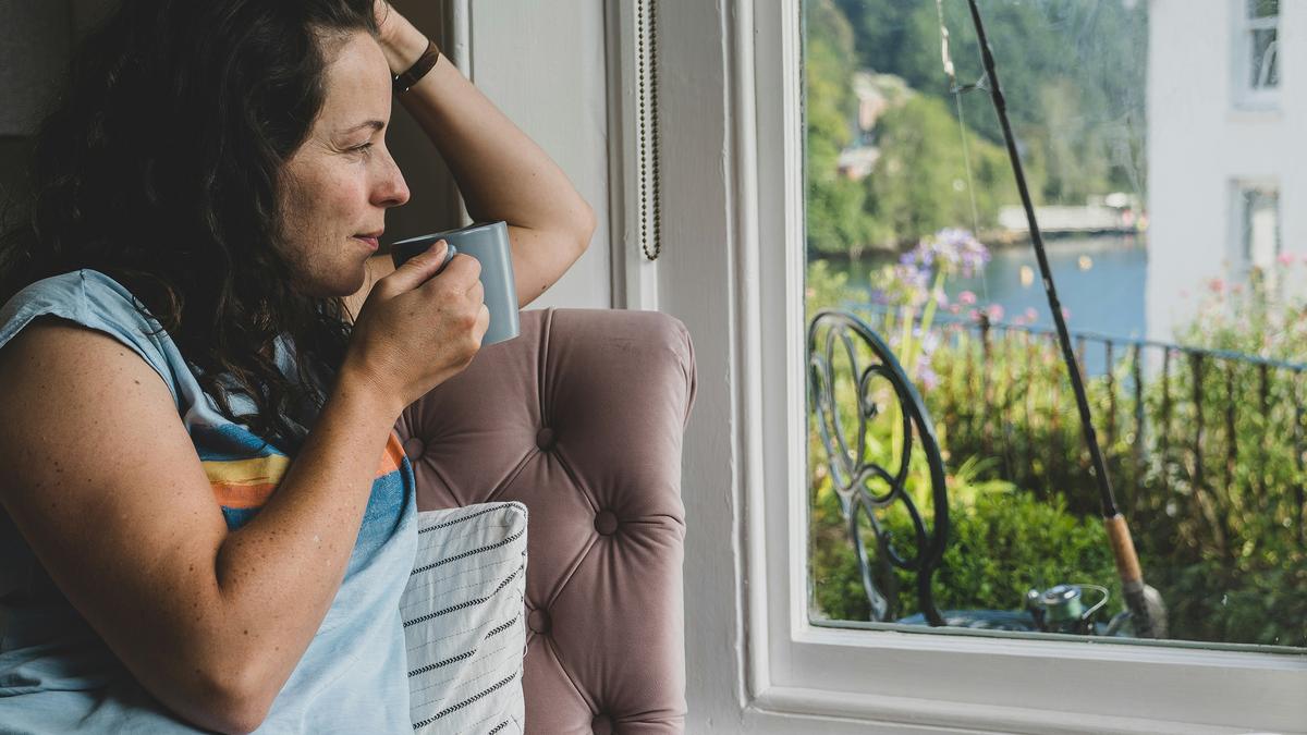 Photo of a woman with a mug gazing out a window, seated on a pink chair near a lake with trees in the background.
