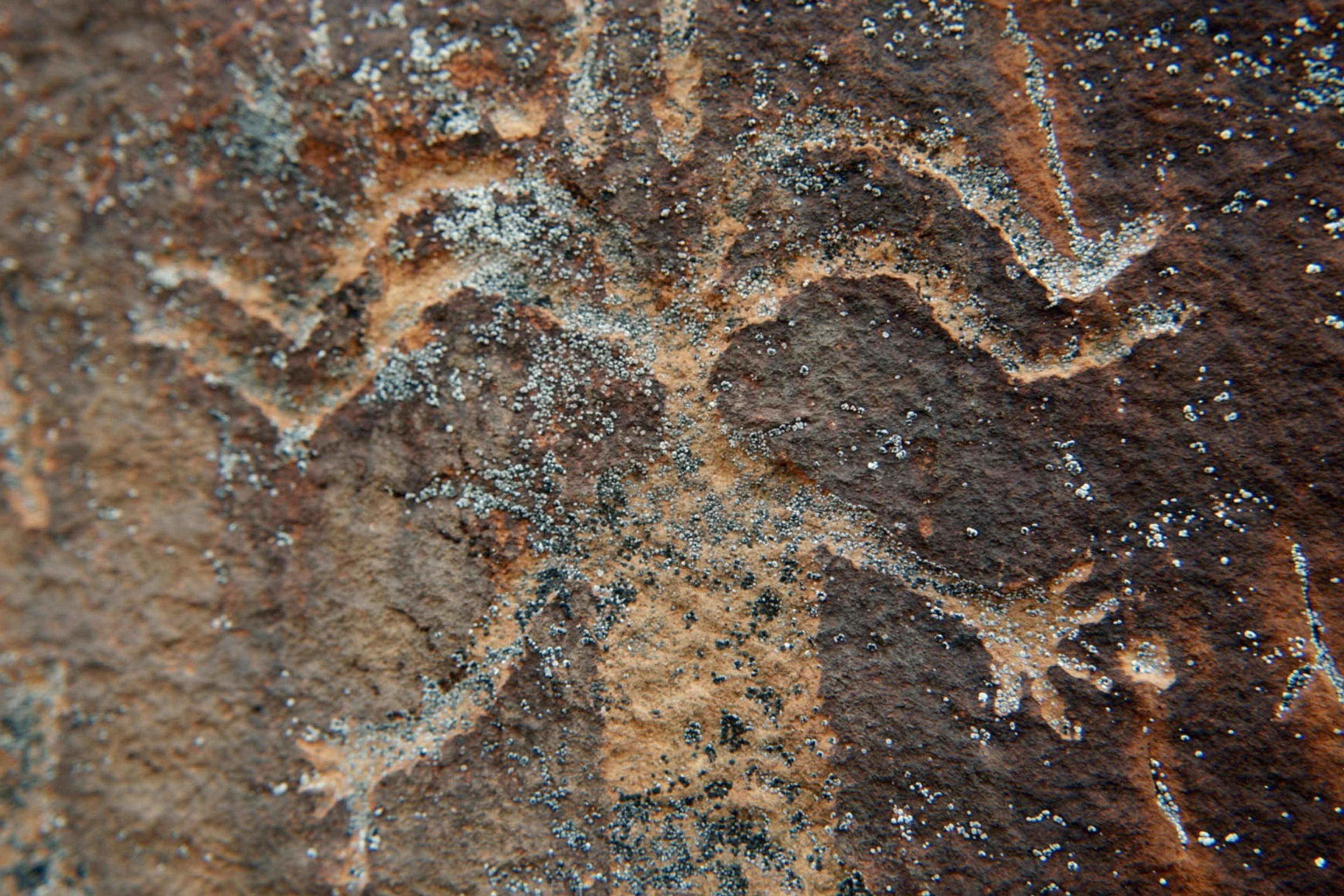 Ancient petroglyph depicting a human-like figure with outstretched arms and legs, etched onto a brown, weathered rock surface.