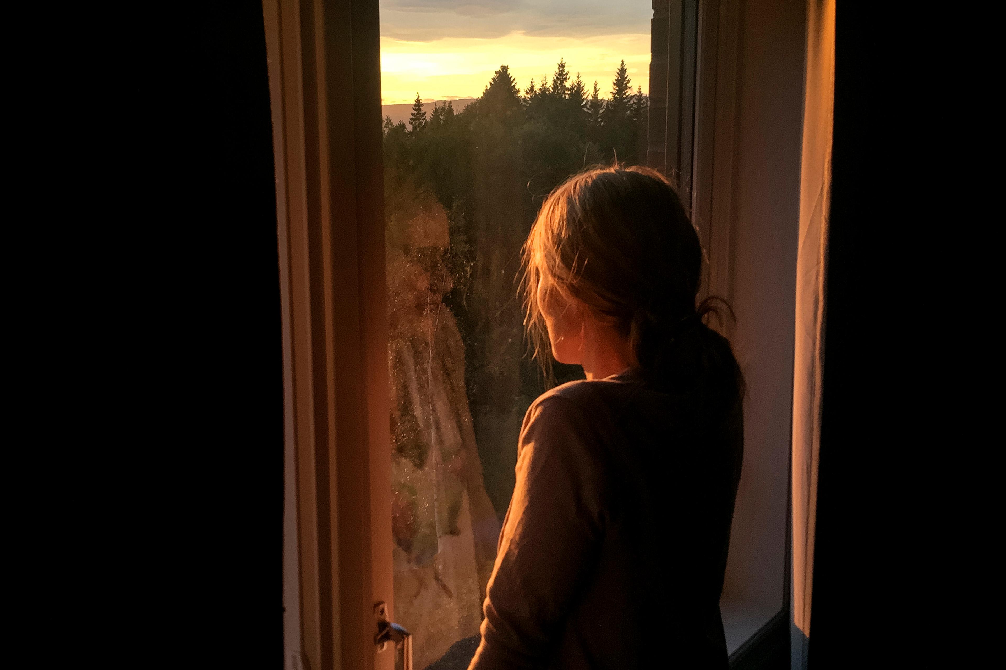 Photo of a person standing by a window watching a forest sunset, with warm light illuminating their silhouette.