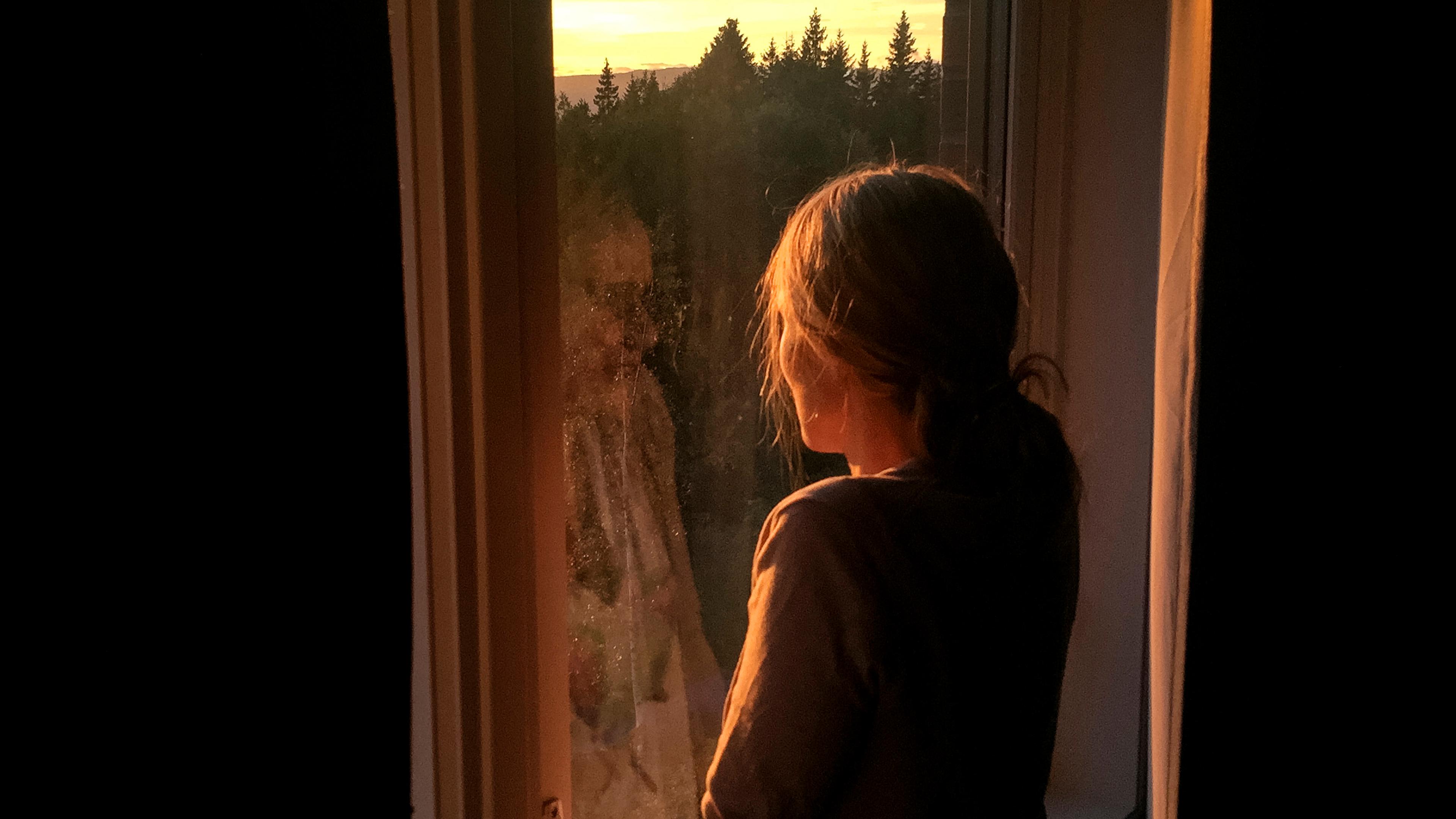 Photo of a person standing by a window watching a forest sunset, with warm light illuminating their silhouette.