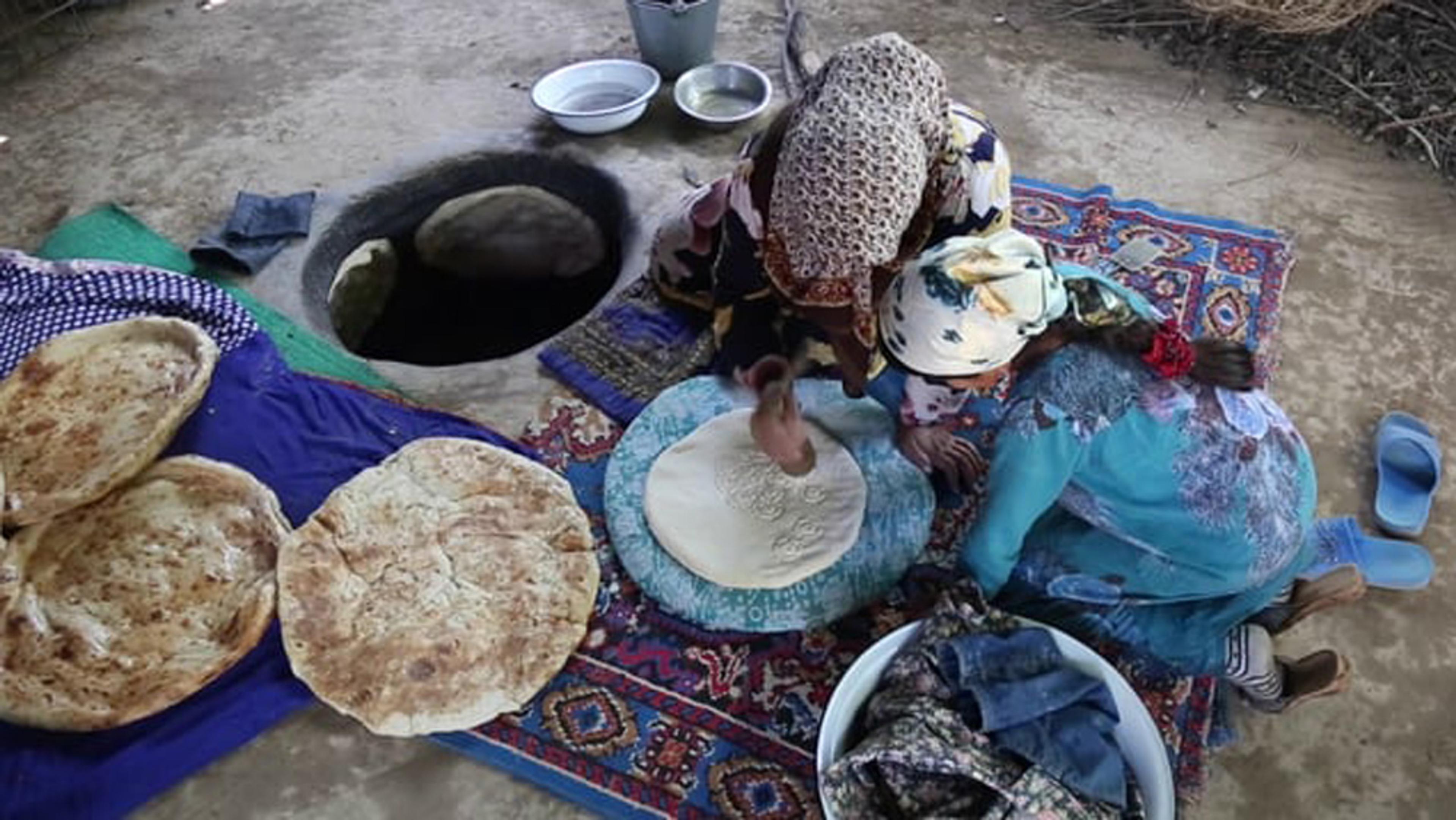 Two women preparing and baking flatbreads on the ground beside a tandoor with rugs, pans and slippers around them.