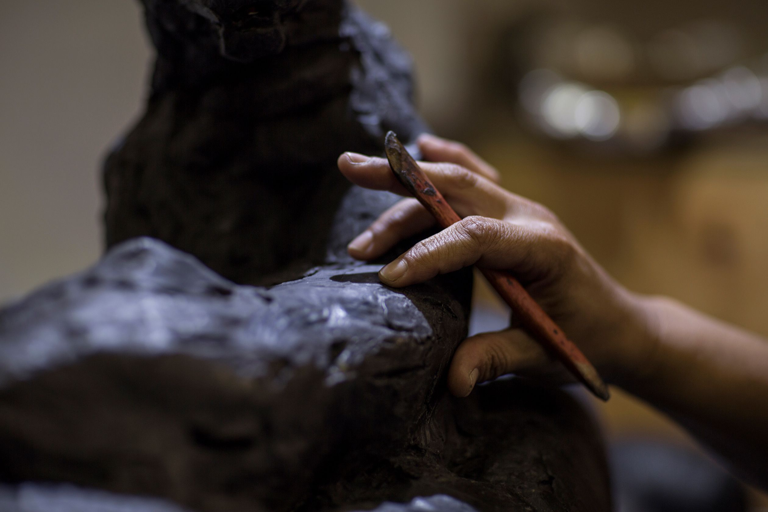 Close-up photo of a sculptor’s hand working clay with a tool, focusing on texture and detail.