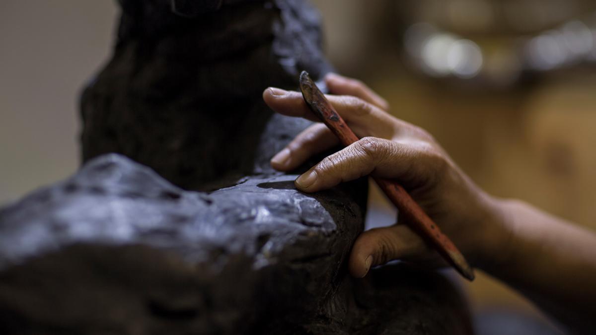 Close-up photo of a sculptor’s hand working clay with a tool, focusing on texture and detail.