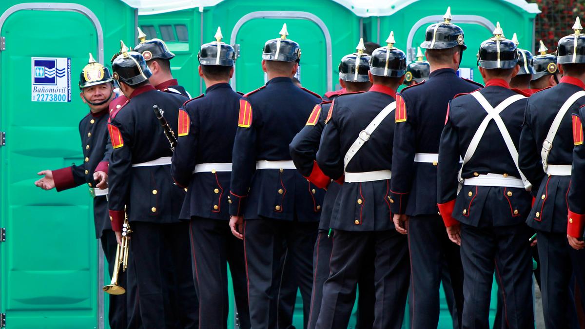 Photo of uniformed men standing outside portable toilets, one gestures invitingly.