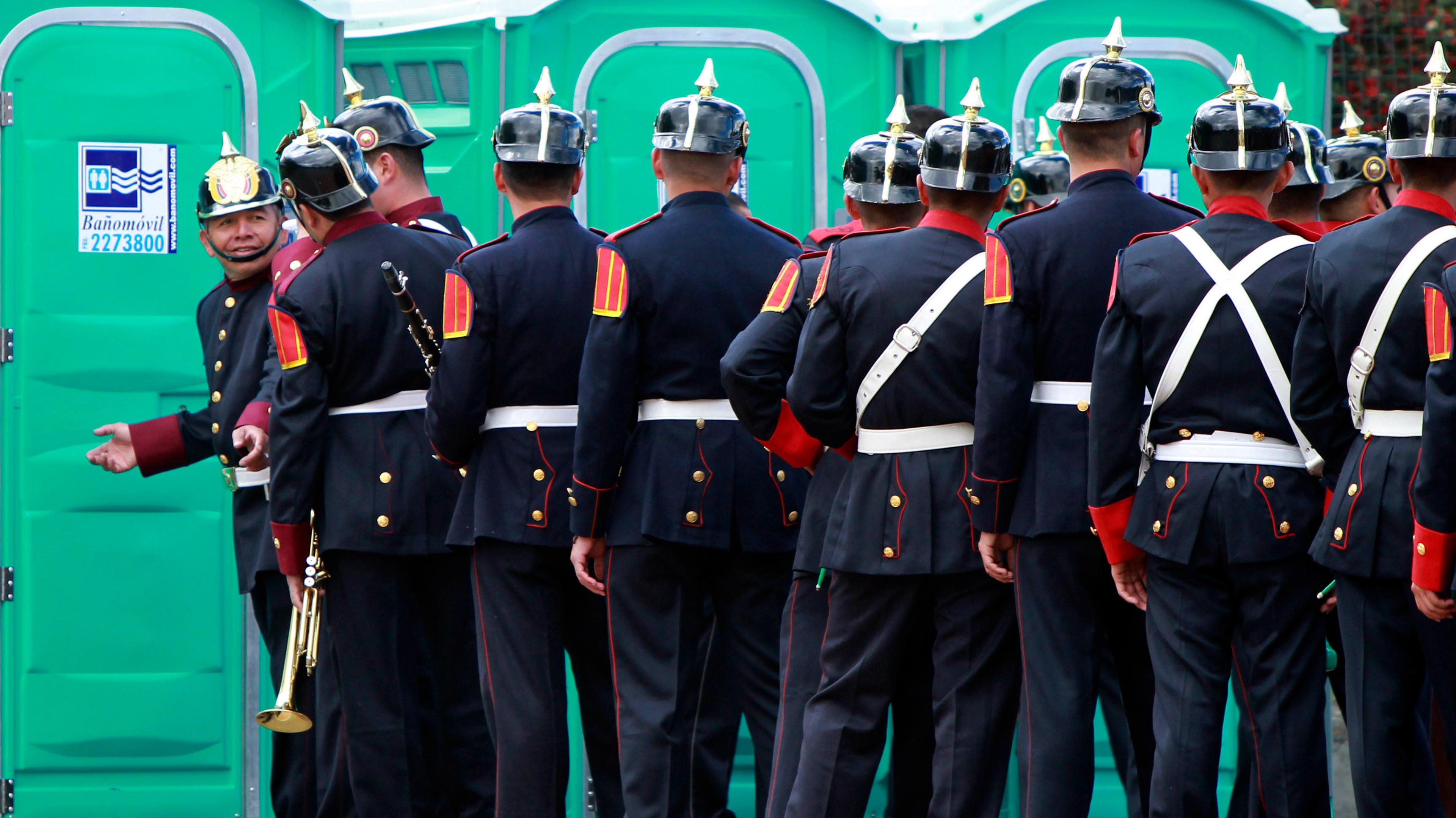 Photo of uniformed men standing outside portable toilets, one gestures invitingly.