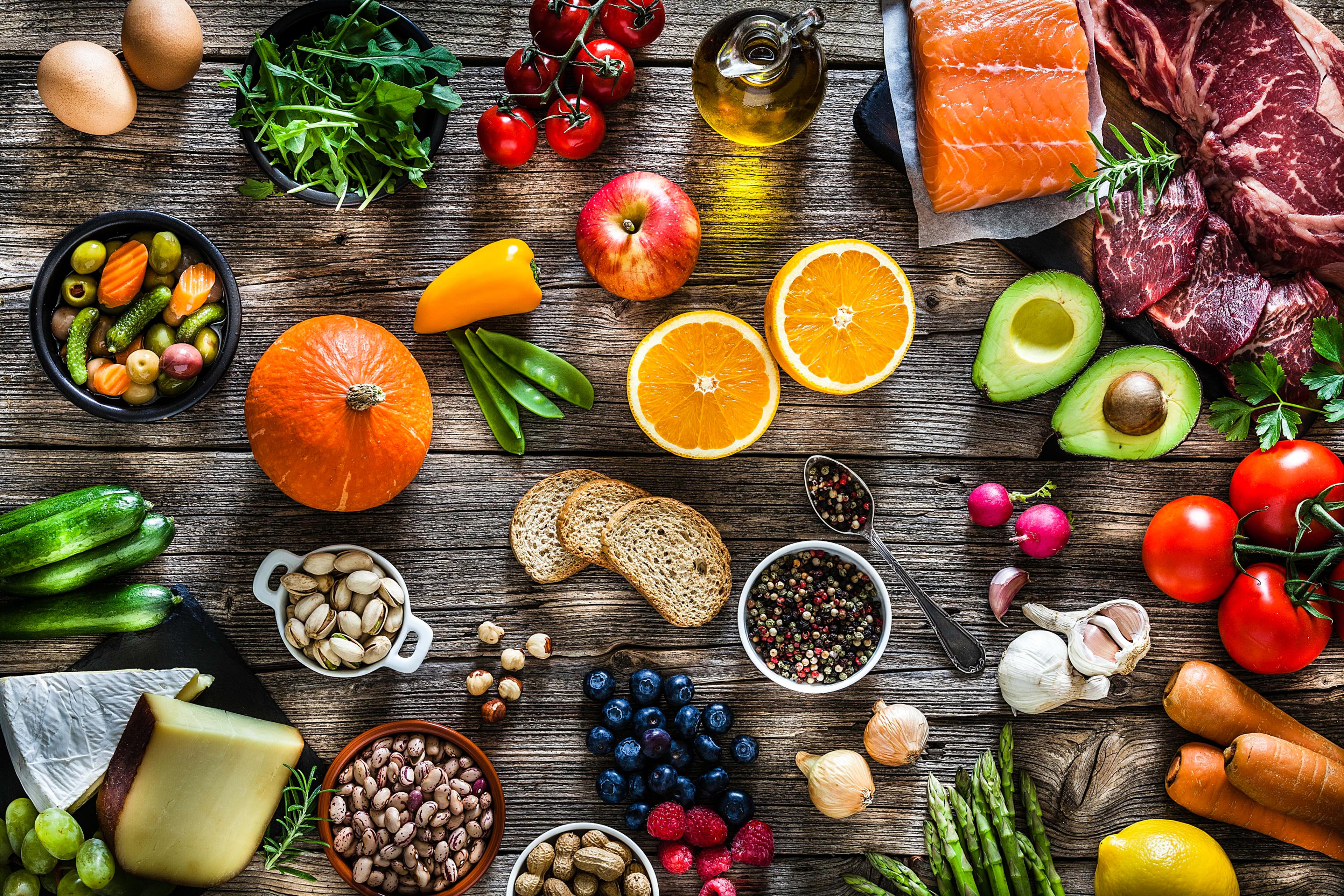 Photo of assorted fresh foods on a wooden table including fruit veg cheese meat fish nuts spices oil bread and herbs.