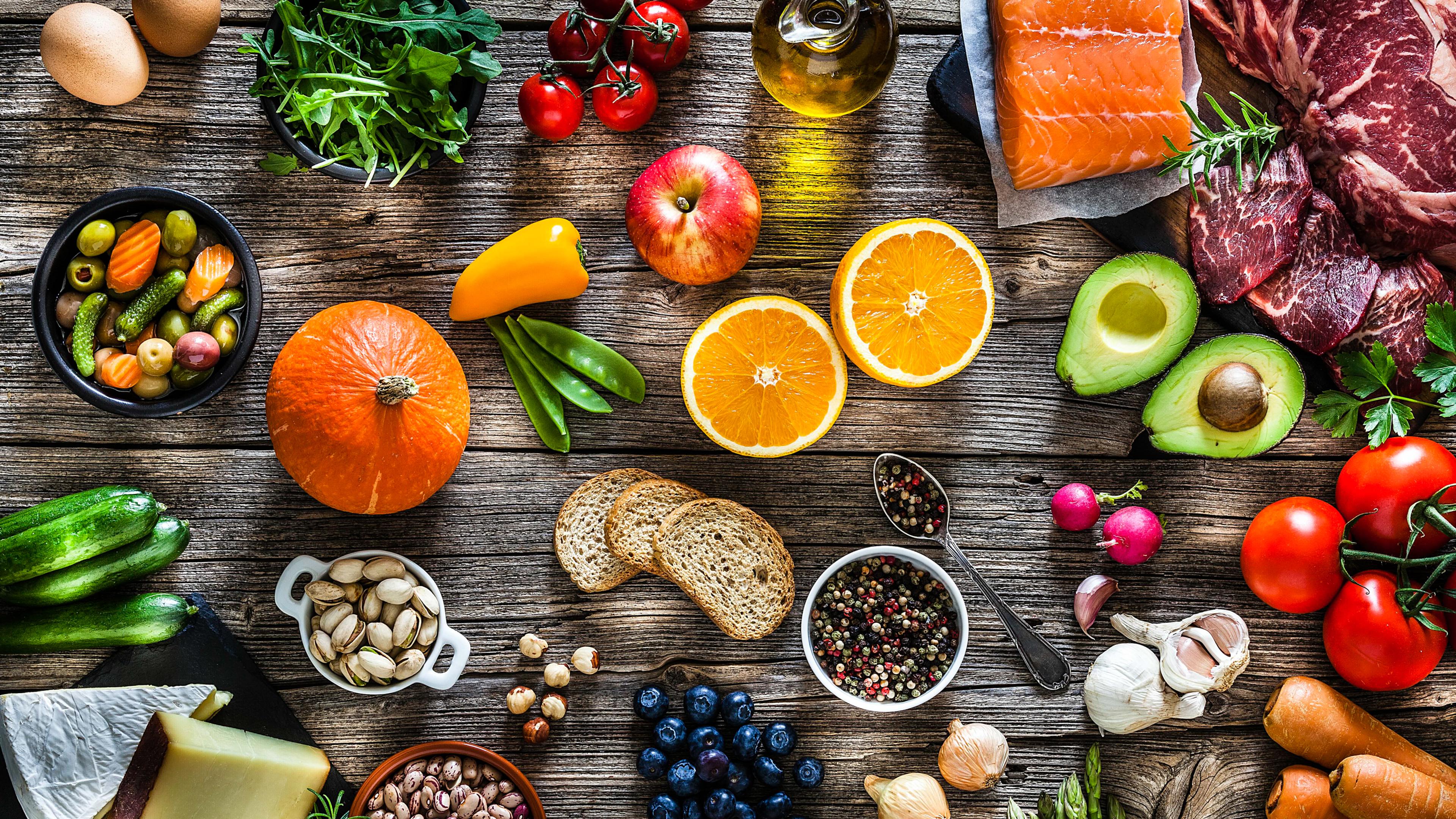 Photo of assorted fresh foods on a wooden table including fruit veg cheese meat fish nuts spices oil bread and herbs.