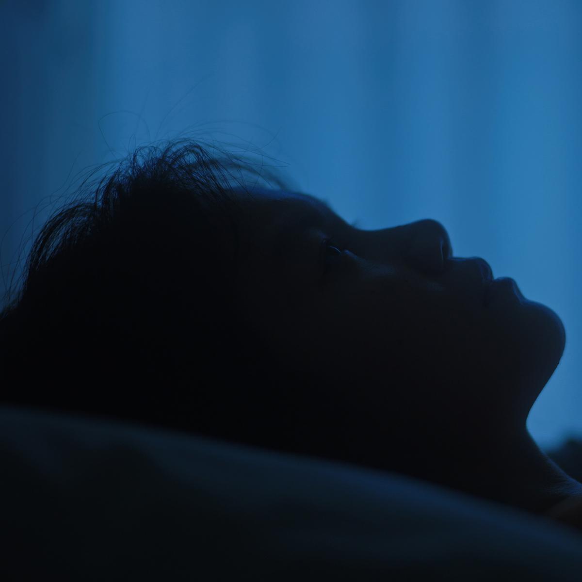 A person lying on their back, silhouetted against a blue background with dim lighting, creating a contemplative mood.