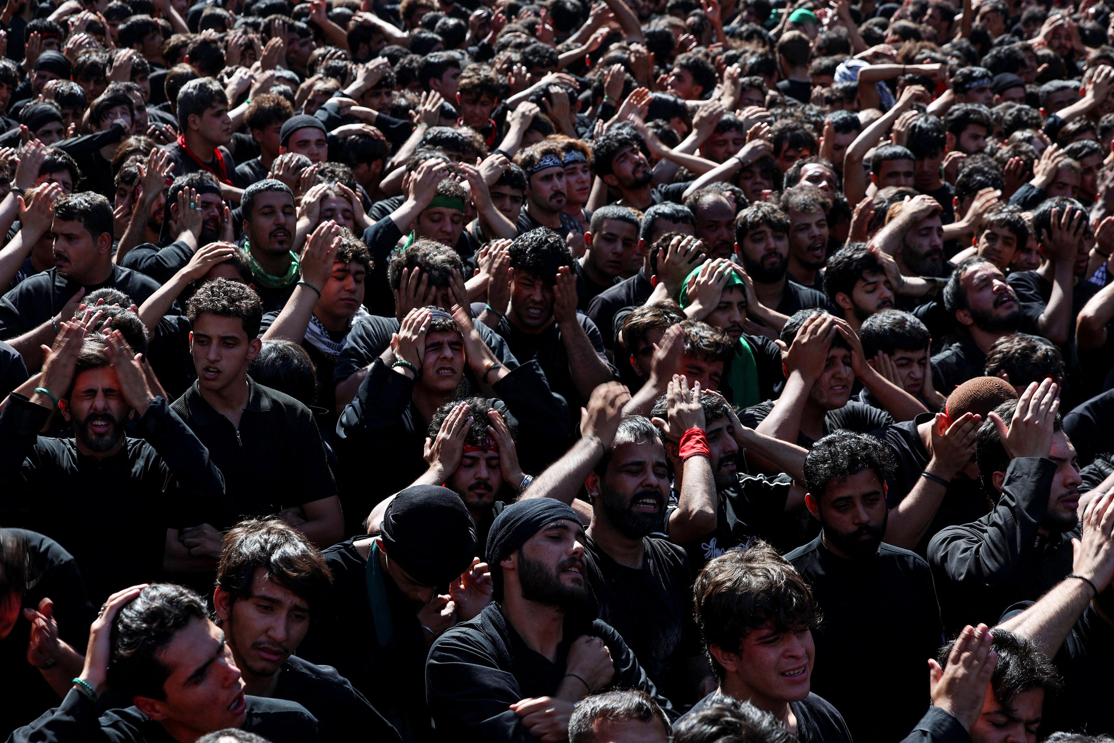 A large crowd of men in black attire participating in a procession with hands on heads, expressing emotion.