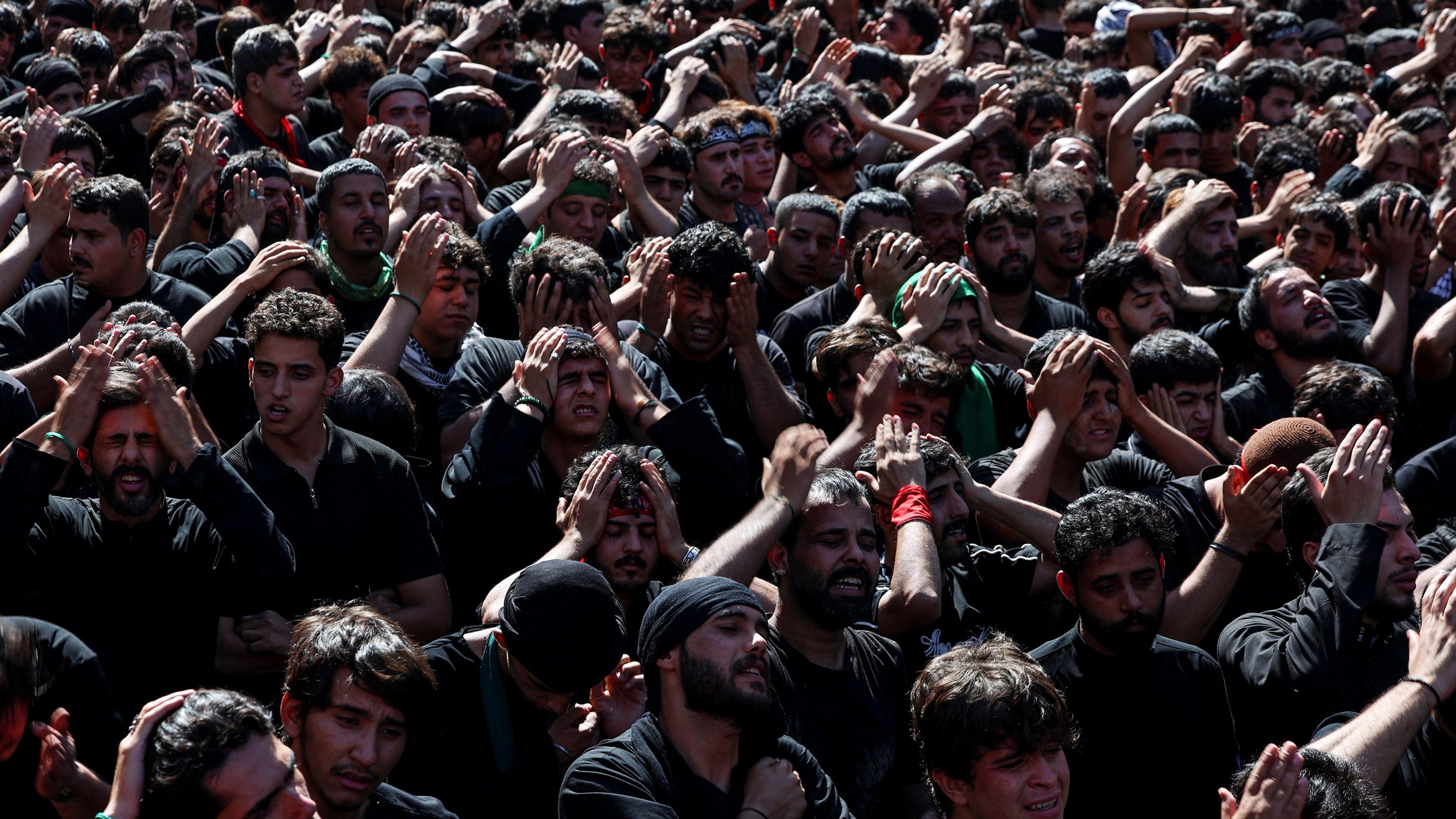 A large crowd of men in black attire participating in a procession with hands on heads, expressing emotion.