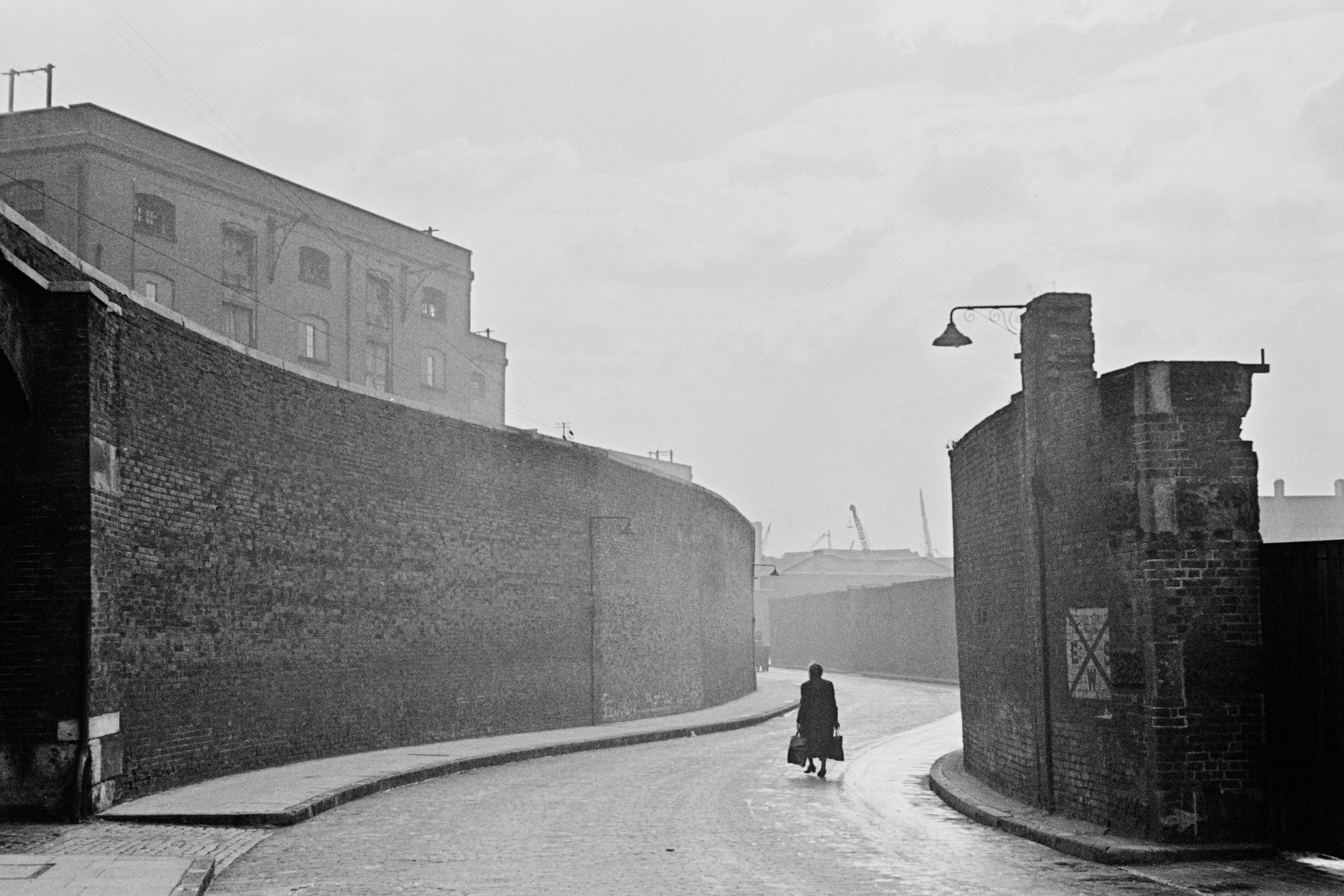 Black and white photo of a solitary figure walking down a misty cobbled street lined by tall brick walls.