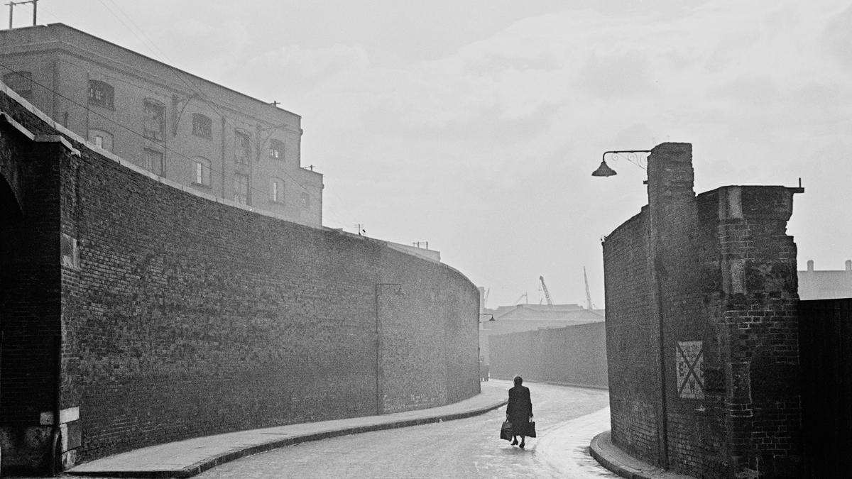 Black and white photo of a solitary figure walking down a misty cobbled street lined by tall brick walls.