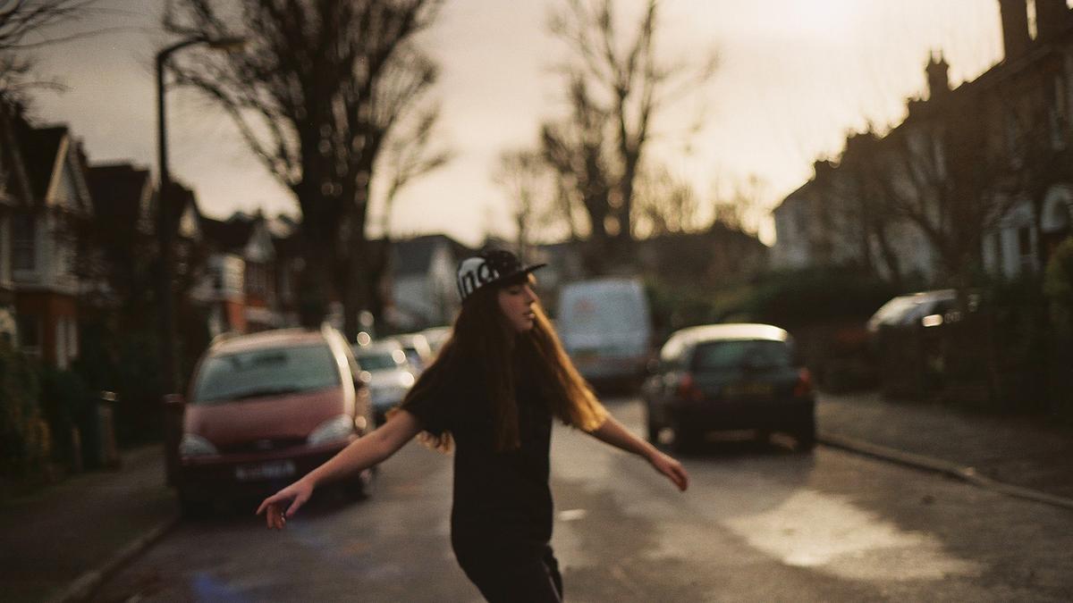 A person in a cap walking on a residential street with cars parked and bare trees in the background.