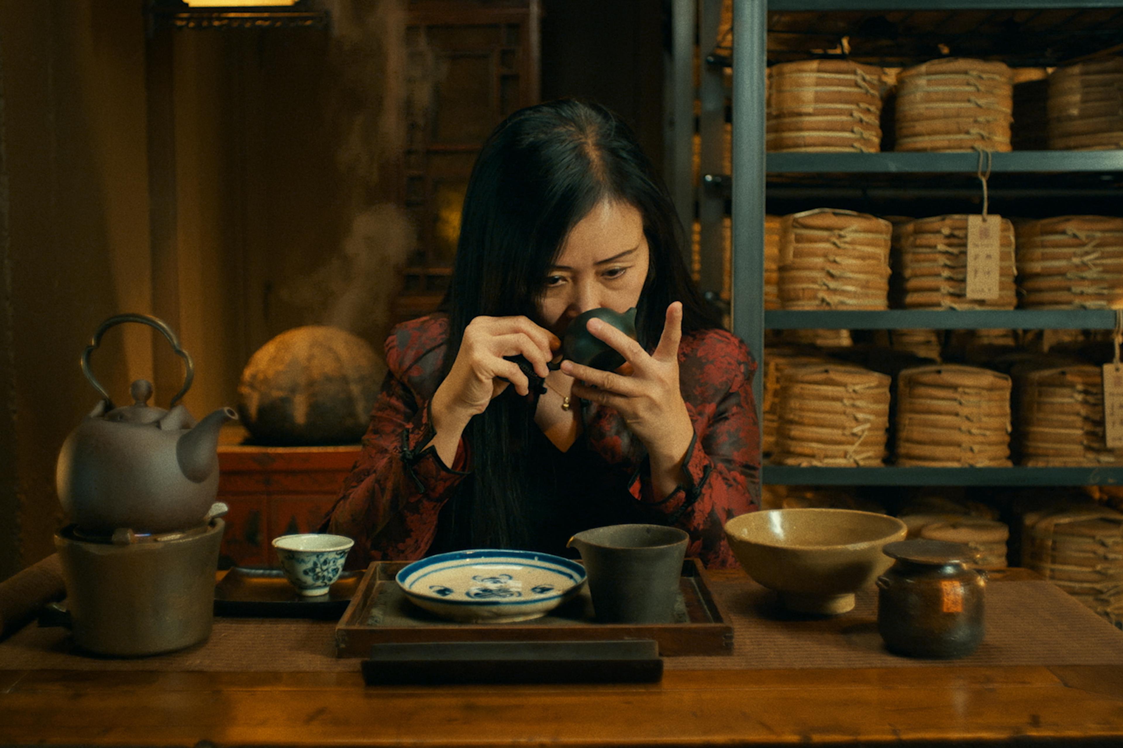 A woman sits at a table with traditional Chinese teacups and teapots. She takes in the aroma of a cup of tea. Behind her are shelves of cylindrical bricks of Pu Erh tea.