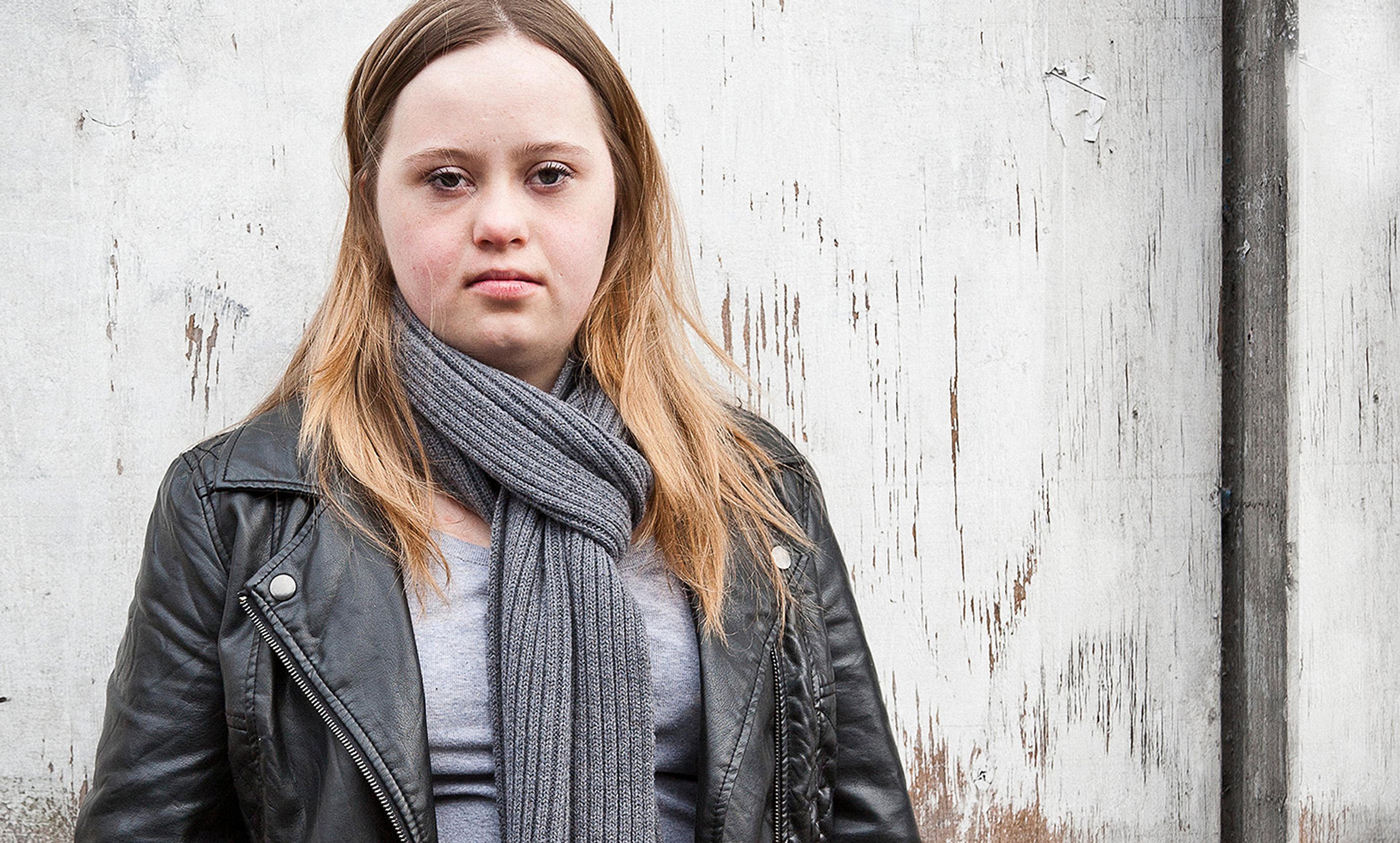A woman with long blonde hair wearing a black leather jacket and grey scarf standing against a weathered wooden wall.