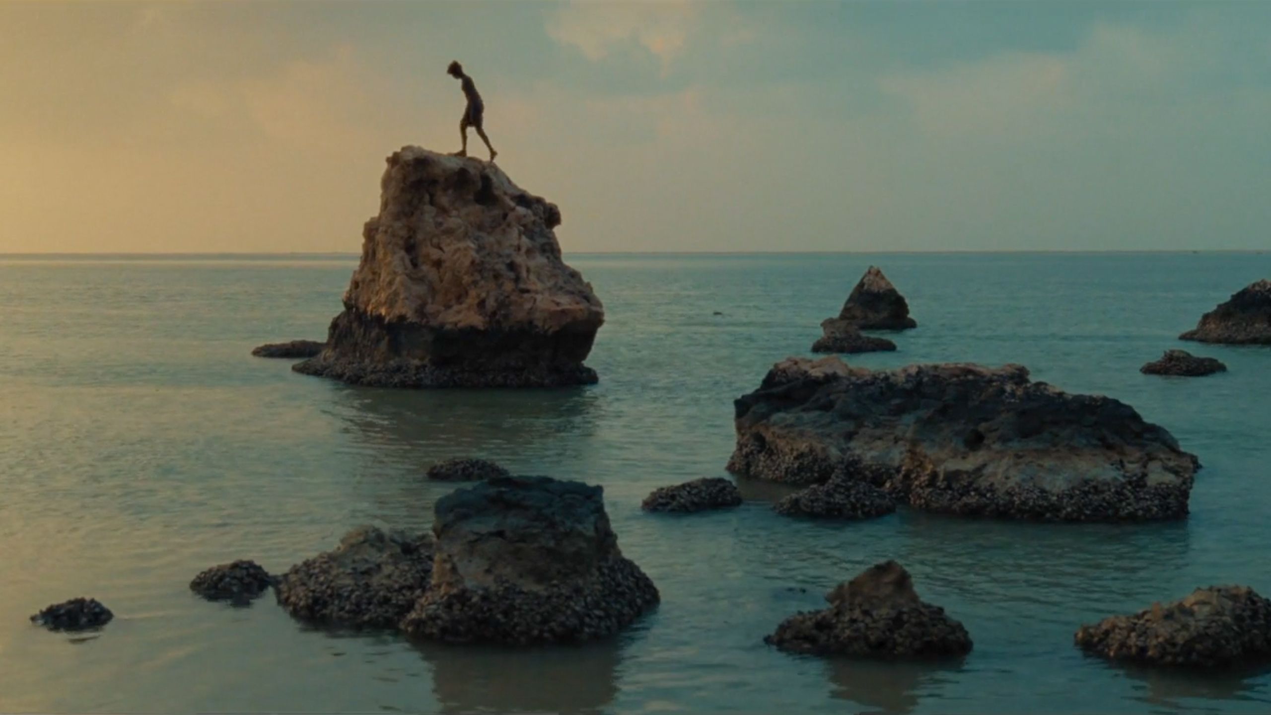 Photo of a person standing on a large rock in the sea during a sunset with scattered smaller rocks around.