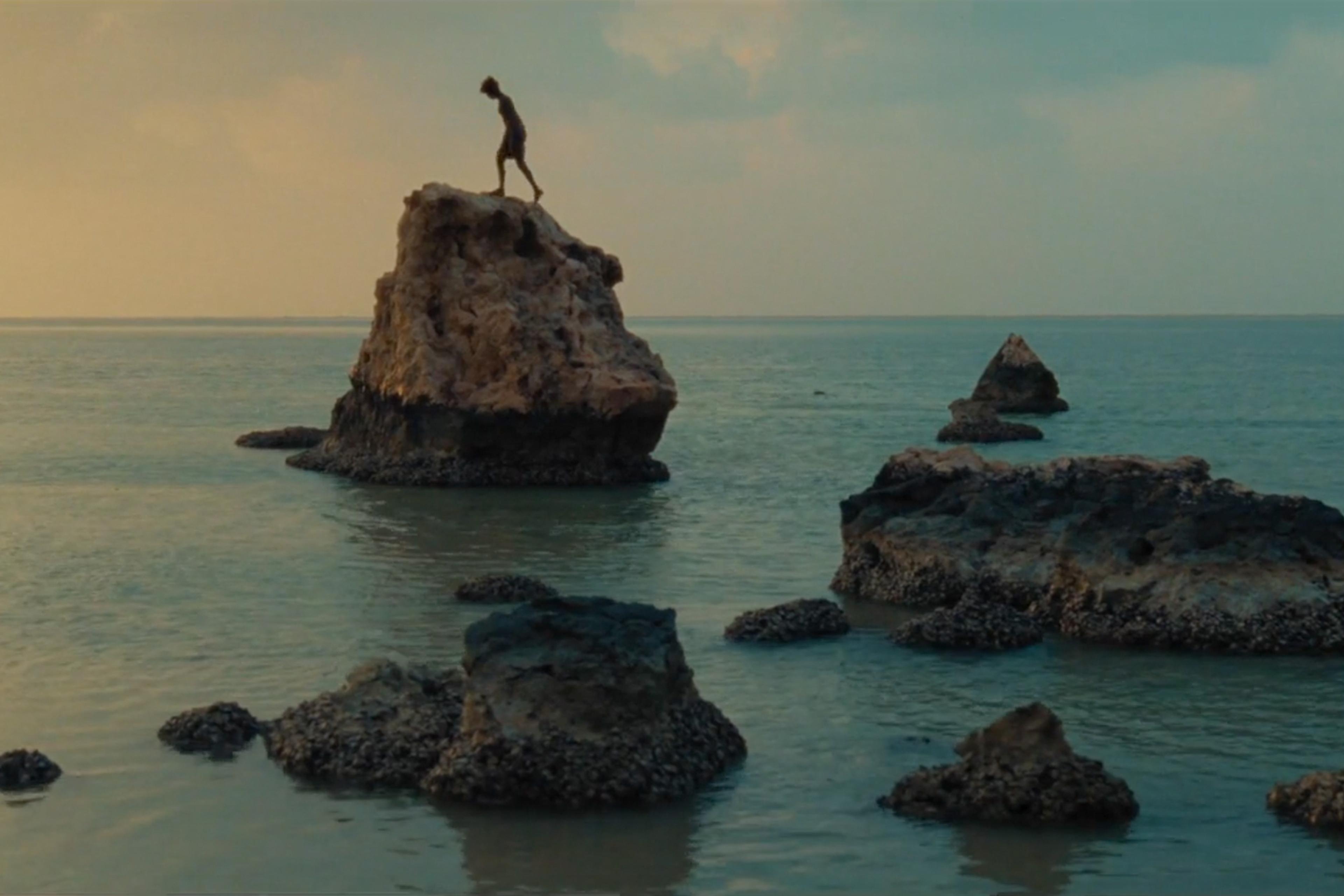 Photo of a person standing on a large rock in the sea during a sunset with scattered smaller rocks around.
