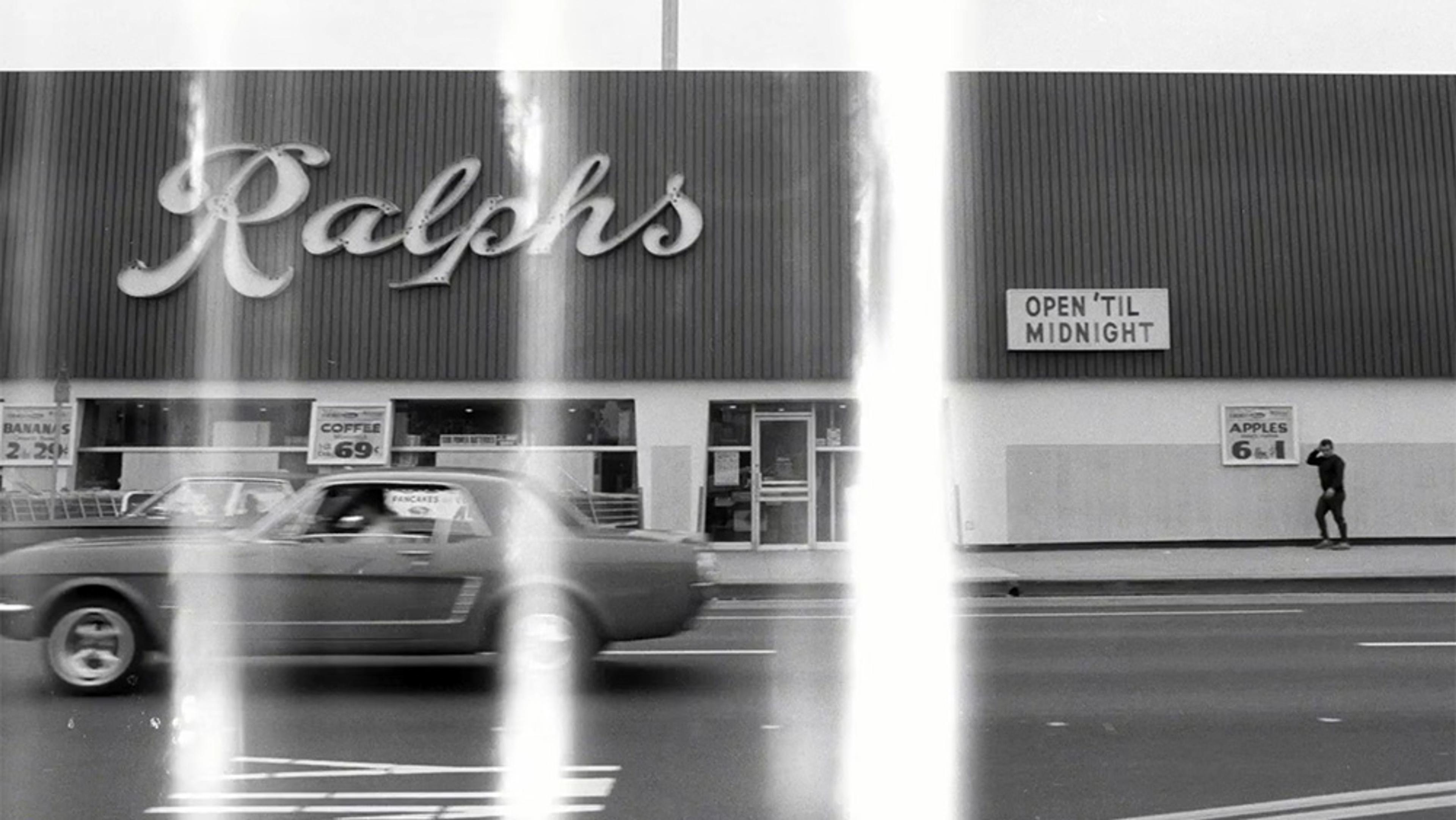 Black-and-white vintage photo of Ralph’s storefront with a car passing by and a person walking, vertical light streaks distort the image.