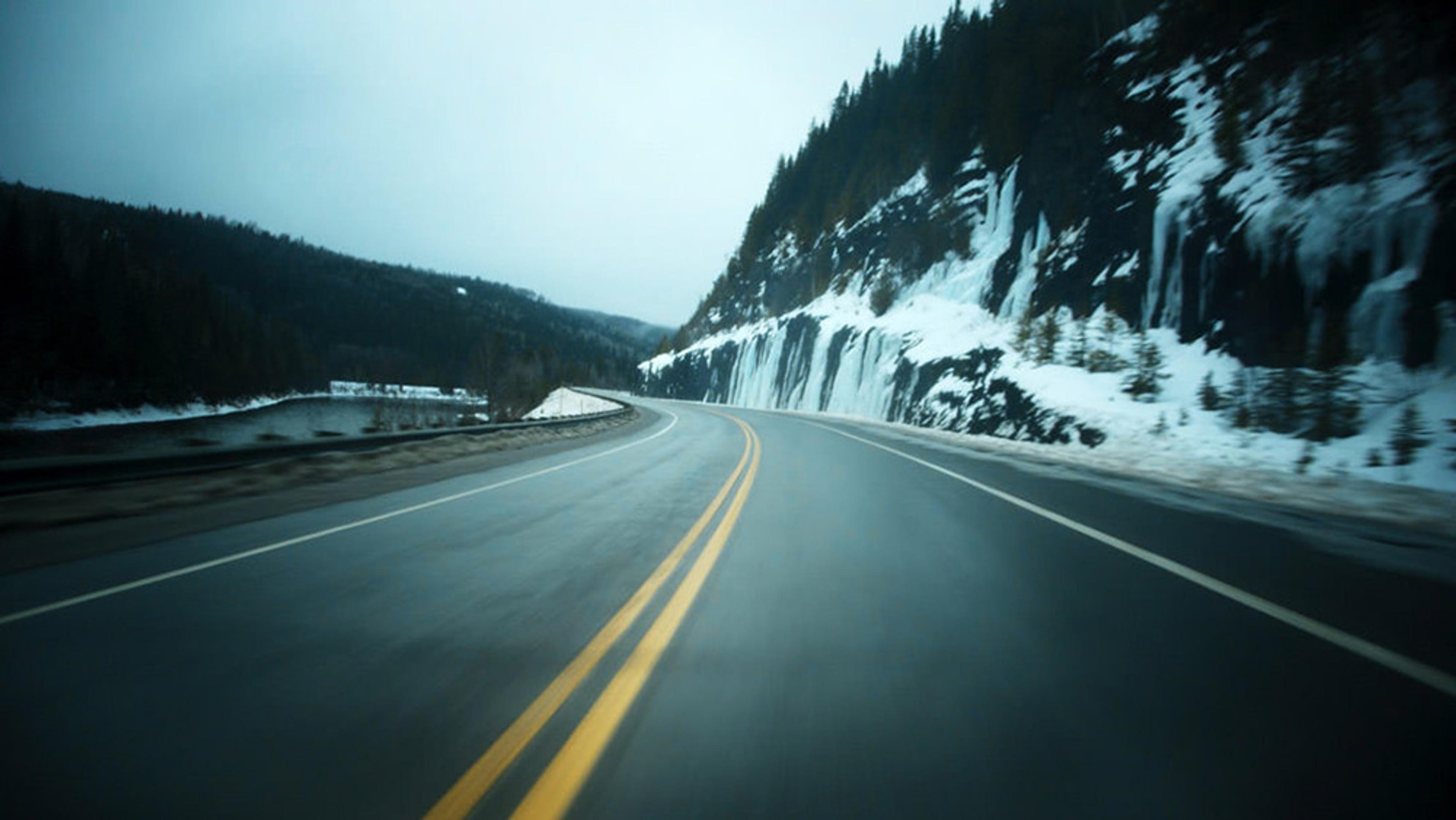 A wet, winding road with yellow lines, flanked by a snowy, wooded hill on one side and a river on the other, under an overcast sky.
