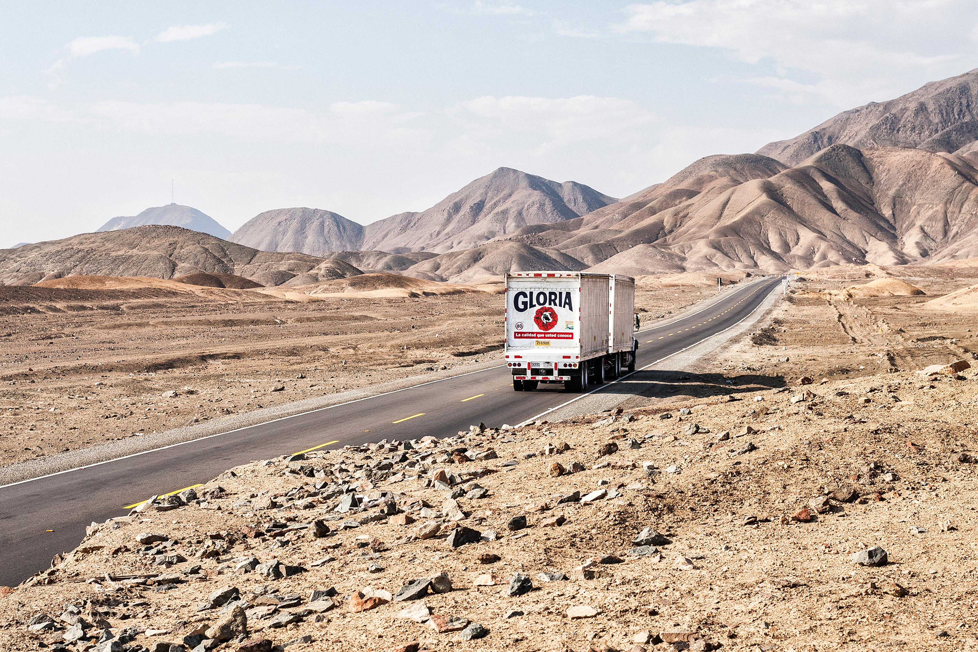 A lorry on a long desert road with rocky terrain and mountains in the background under a partly cloudy sky.