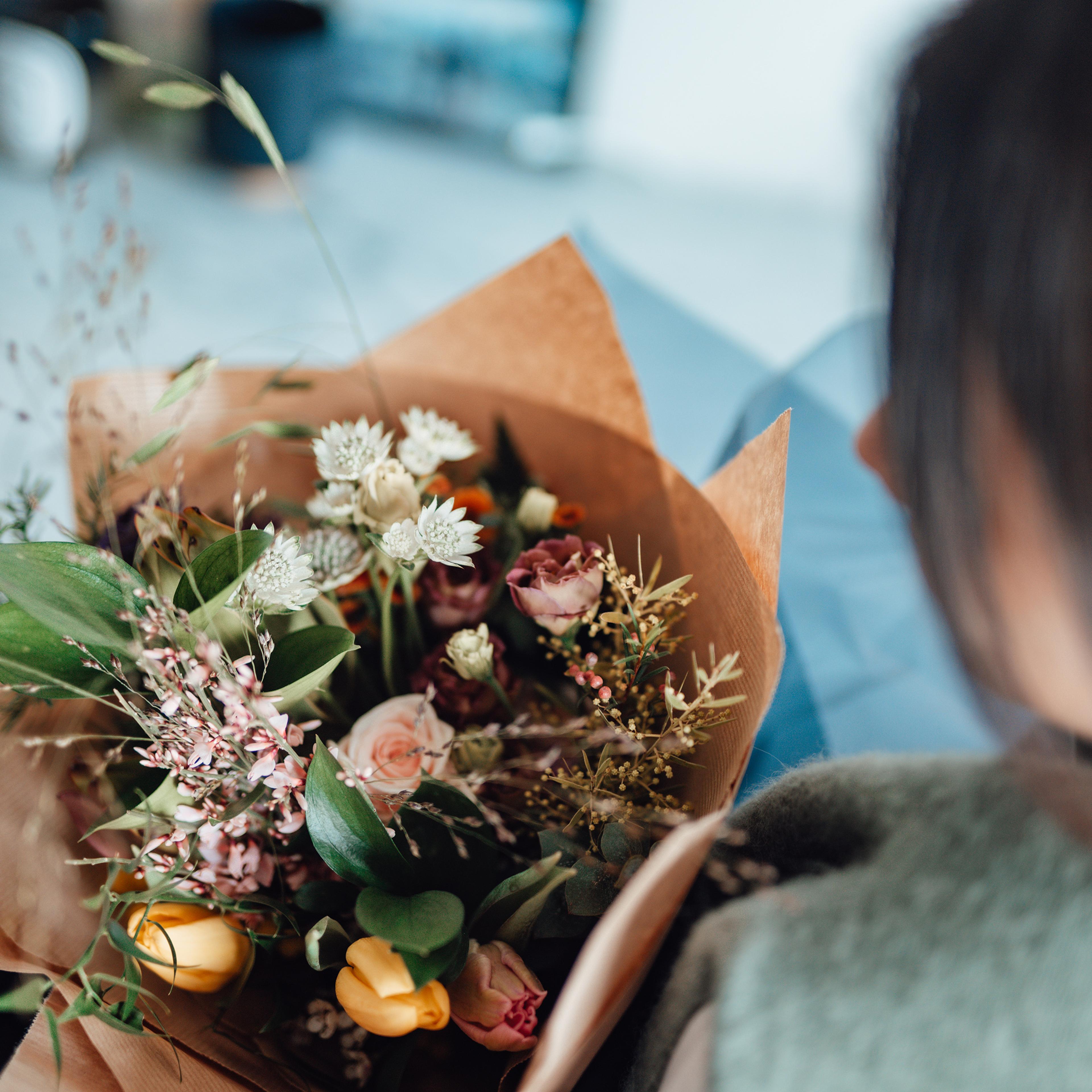 A photo showing a person holding a bouquet of mixed flowers wrapped in brown paper, viewed from above and behind.