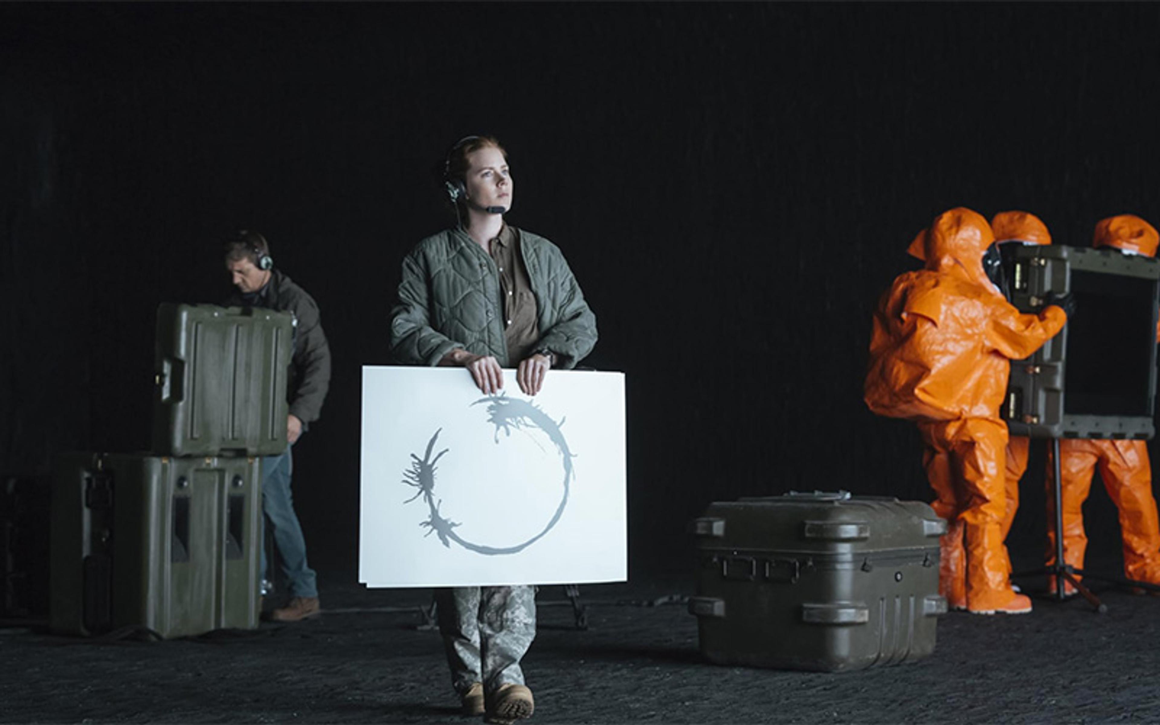 A woman holding a paper card displaying a circular symbol, with people in orange hazmat suits handling equipment in the background.