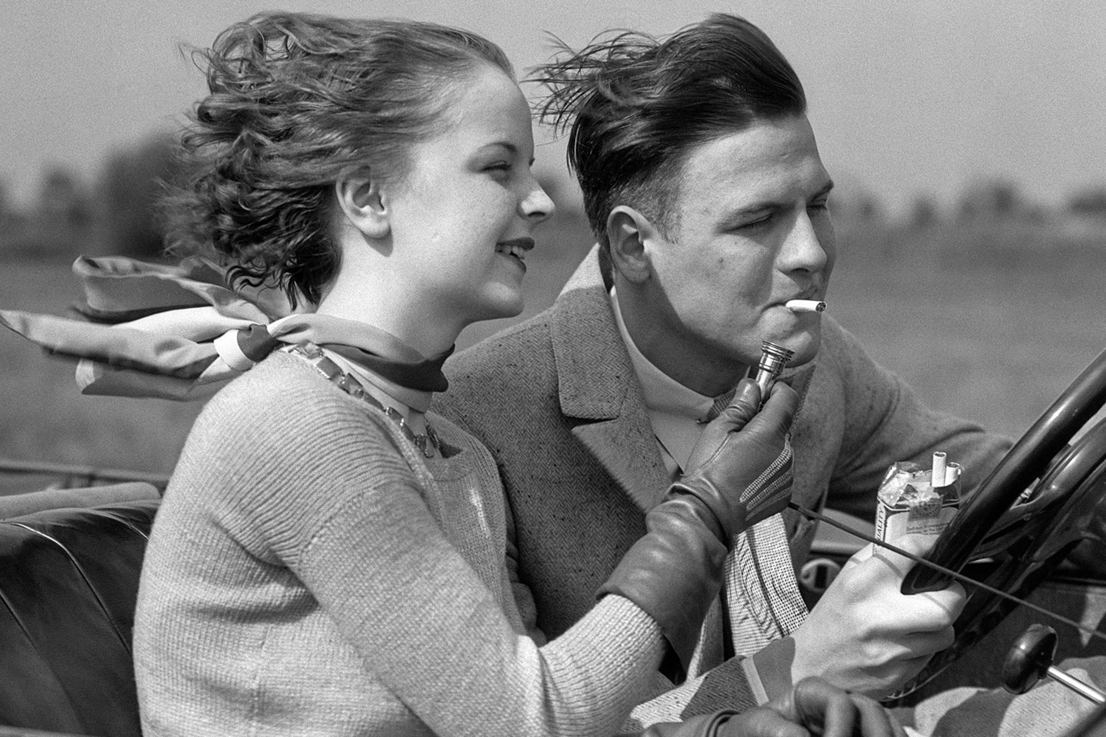 A couple in vintage attire driving a convertible, with the woman lighting the man’s cigarette. They appear to be enjoying a breezy day.