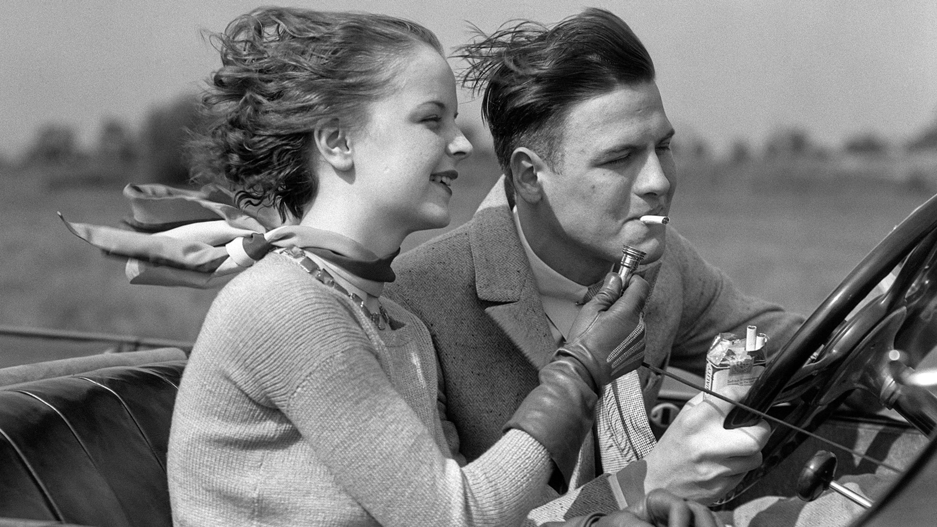 A couple in vintage attire driving a convertible, with the woman lighting the man’s cigarette. They appear to be enjoying a breezy day.