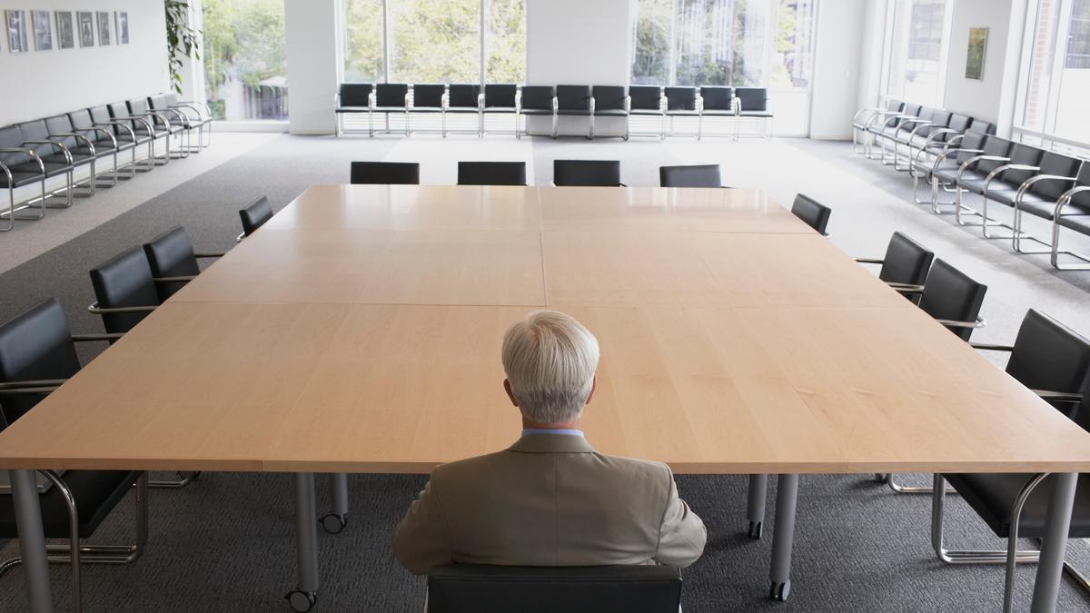 A man with white hair in a suit sitting at the head of an empty boardroom table, facing forward, back toward the camera. Dozens of empty chairs line the outer edges of the room; windows at the rear of the room show trees outside.