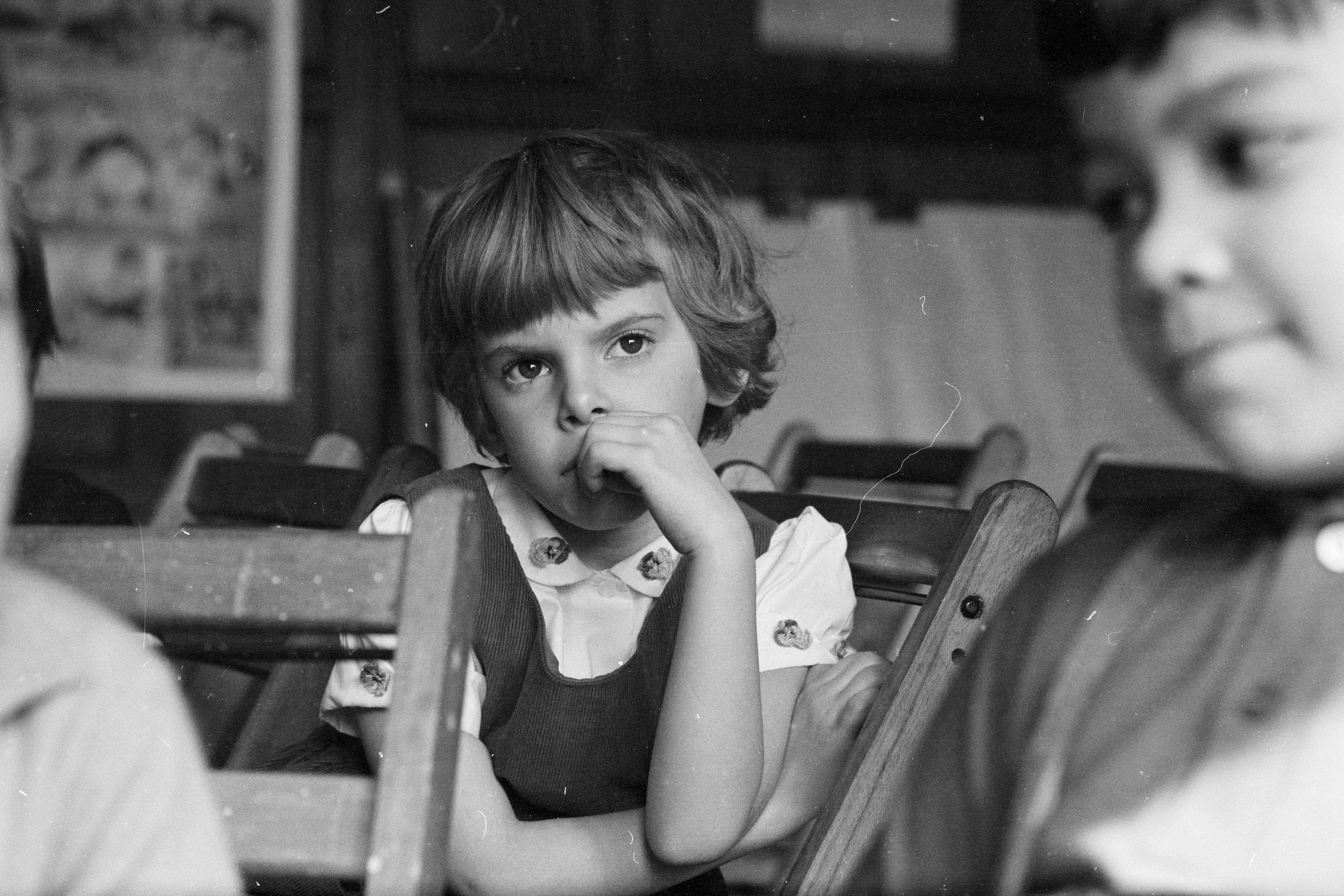 Black and white photo of a thoughtful child in a classroom sitting with hand on chin, other children nearby.