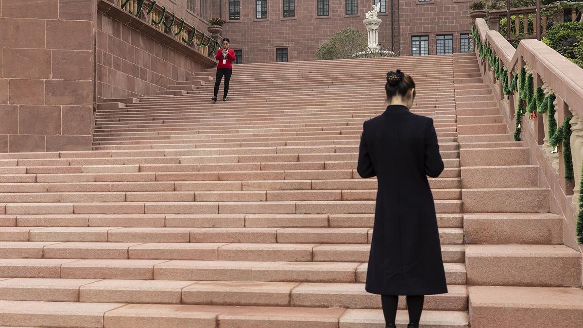 Two people on wide stone steps outside a large building adorned with garlands and a distant statue.