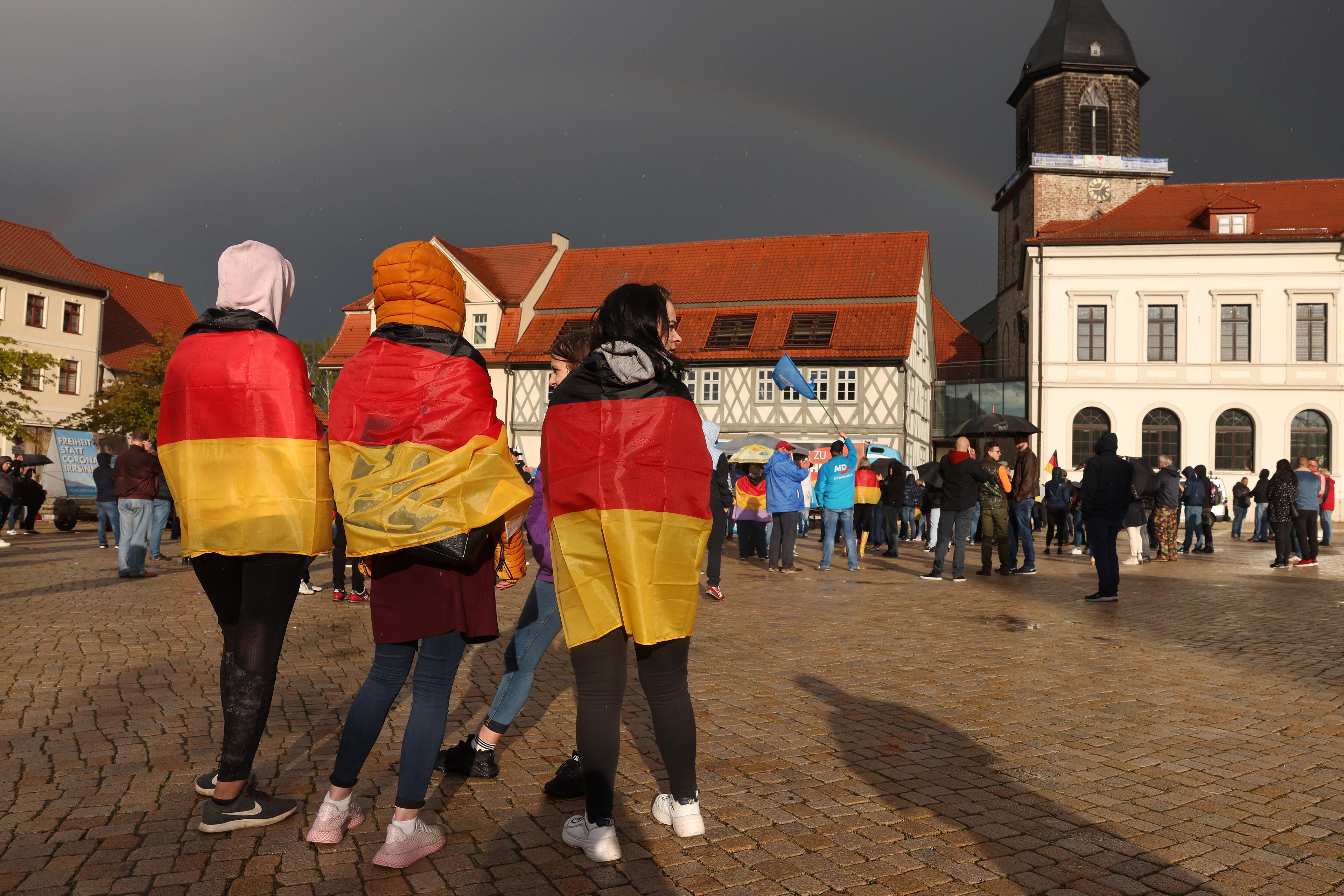 Three people draped in German flags standing in a town square during a gathering, with a rainbow in the cloudy sky.