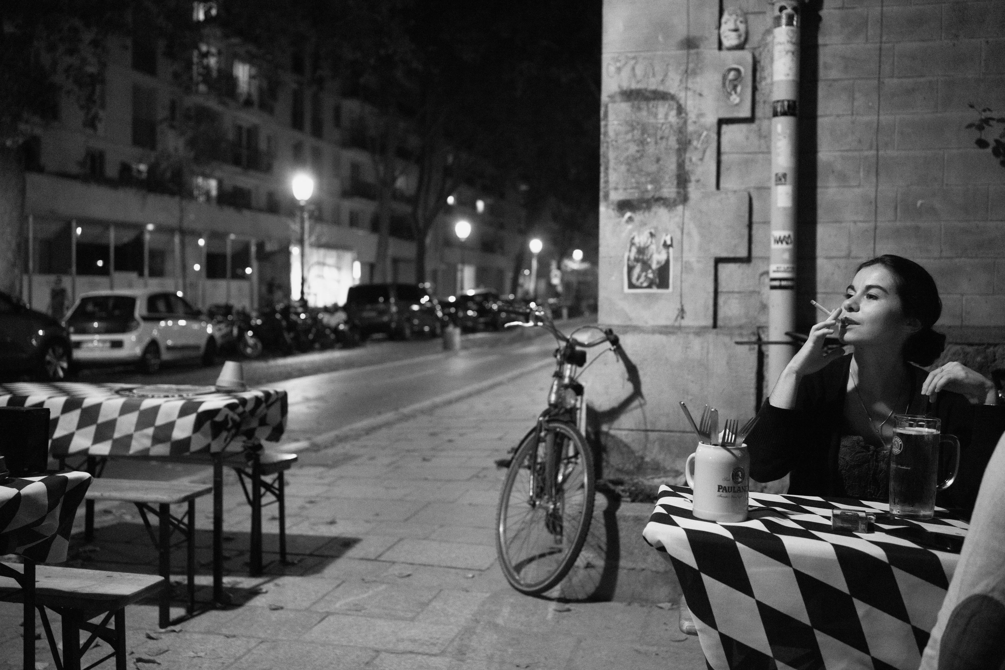 Black-and-white photo of a woman smoking at an outdoor cafe at night with tables, a bicycle and streetlights in the background.