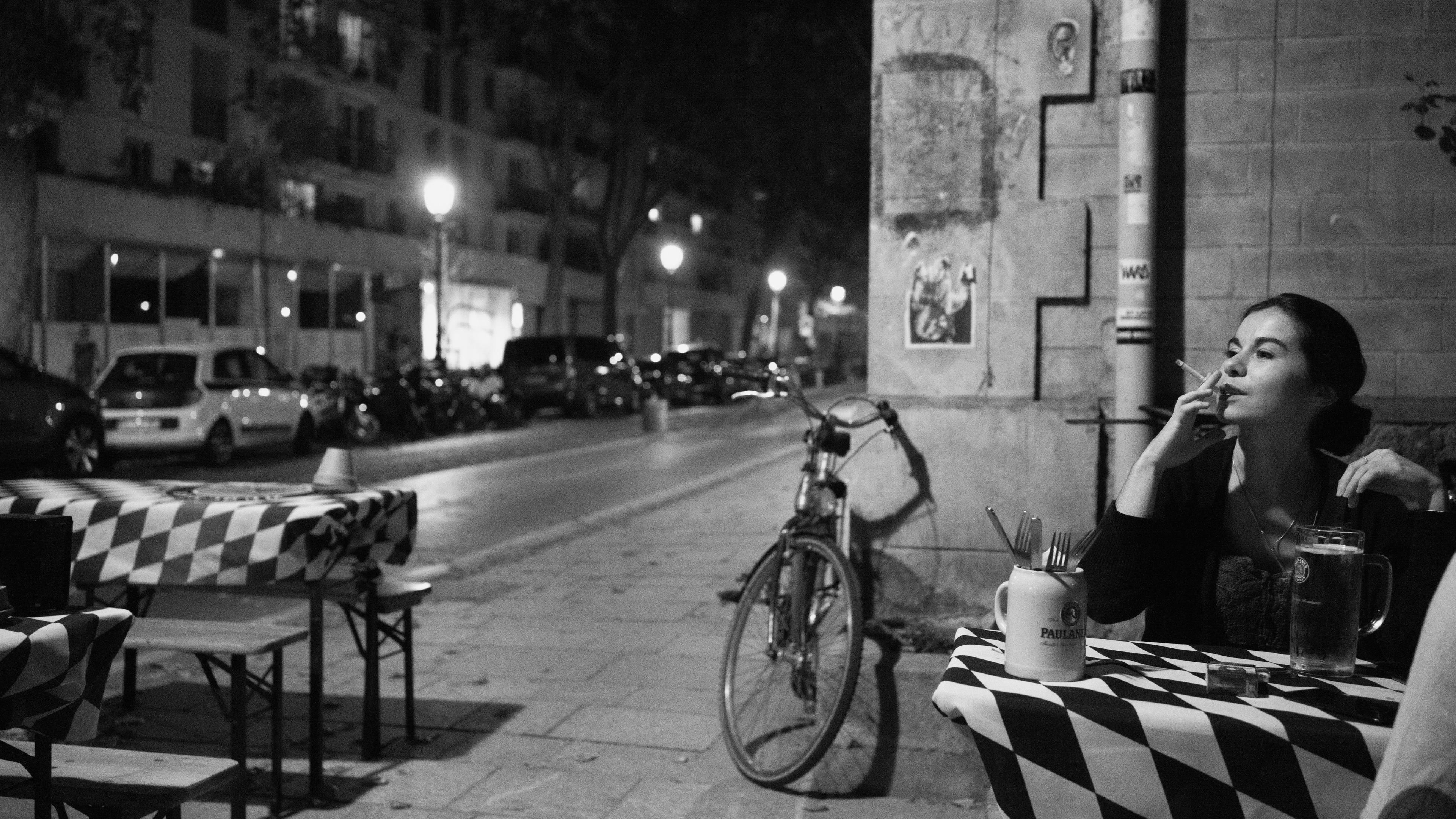 Black-and-white photo of a woman smoking at an outdoor cafe at night with tables, a bicycle and streetlights in the background.