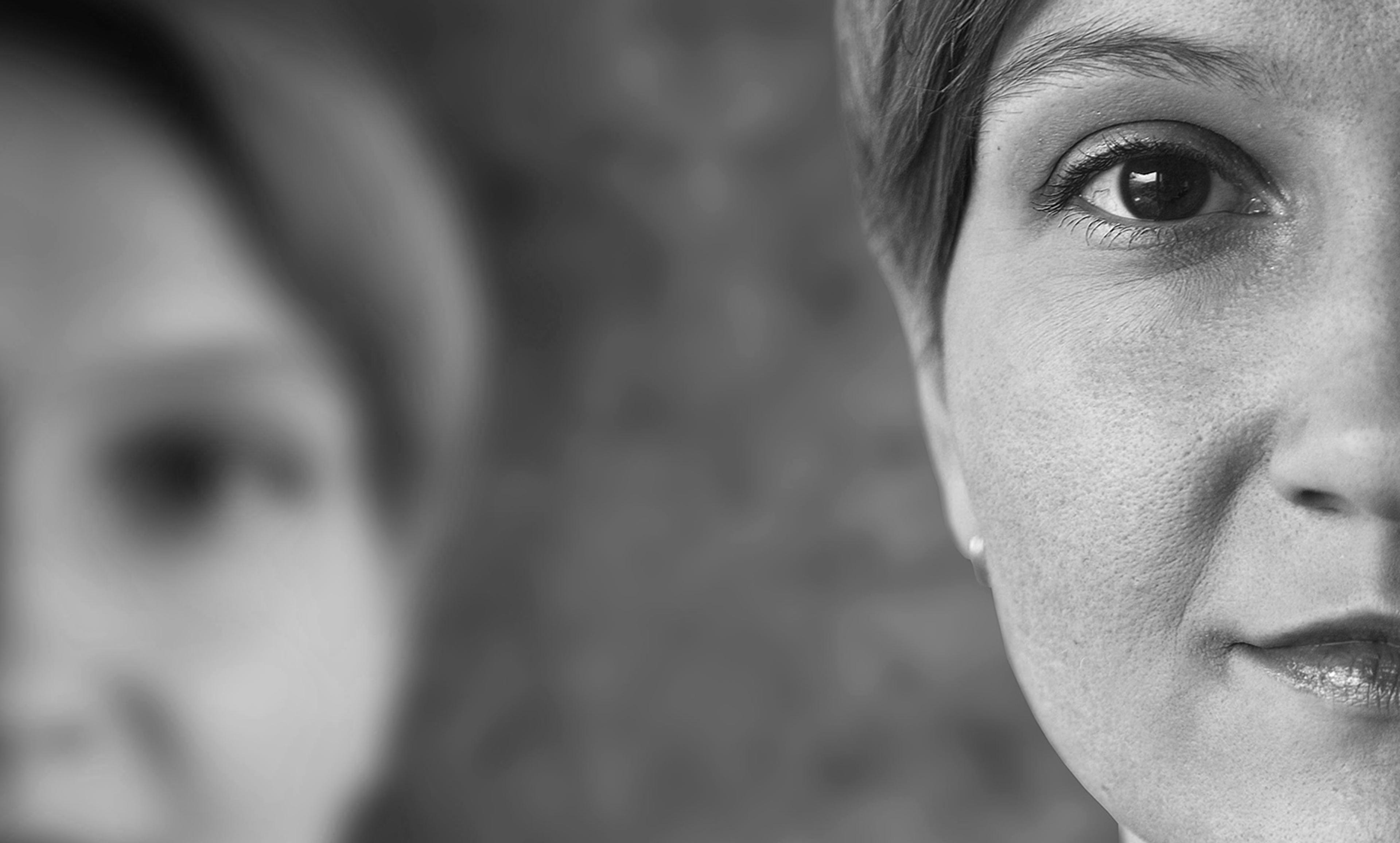 Black and white photo showing half of a woman’s face in focus, with another out-of-focus face in the background.