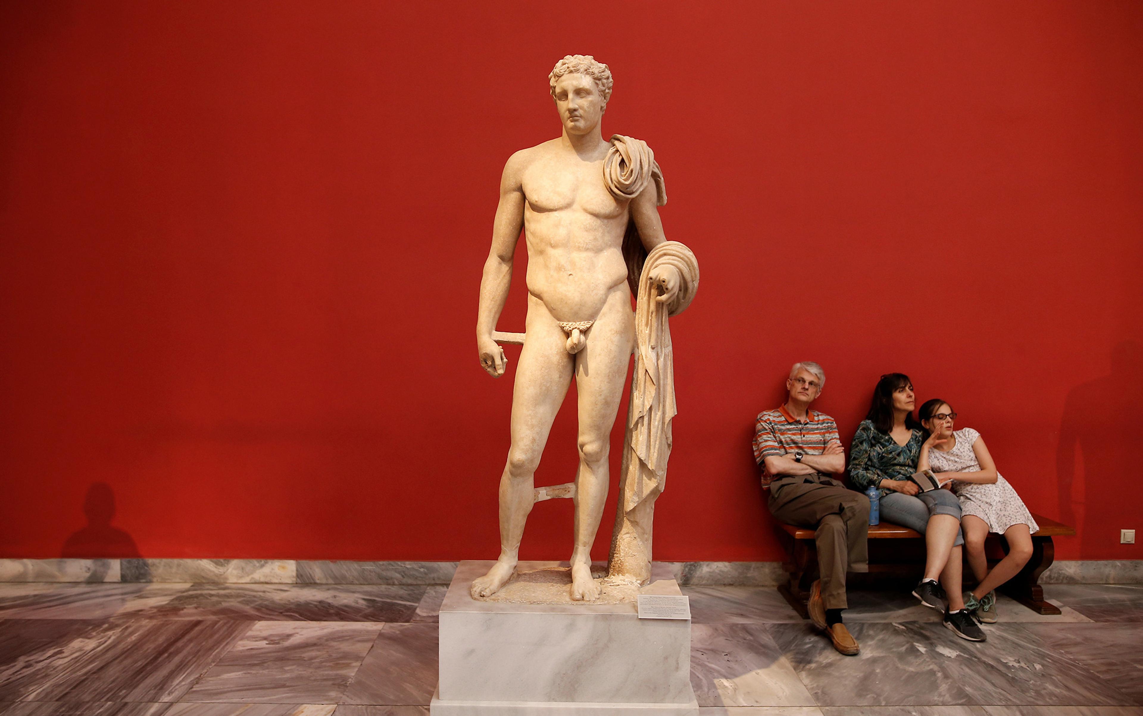 Photo of an ancient male statue with a cloth draped over his shoulder and arm, three people are sitting on a bench to the side of the statue