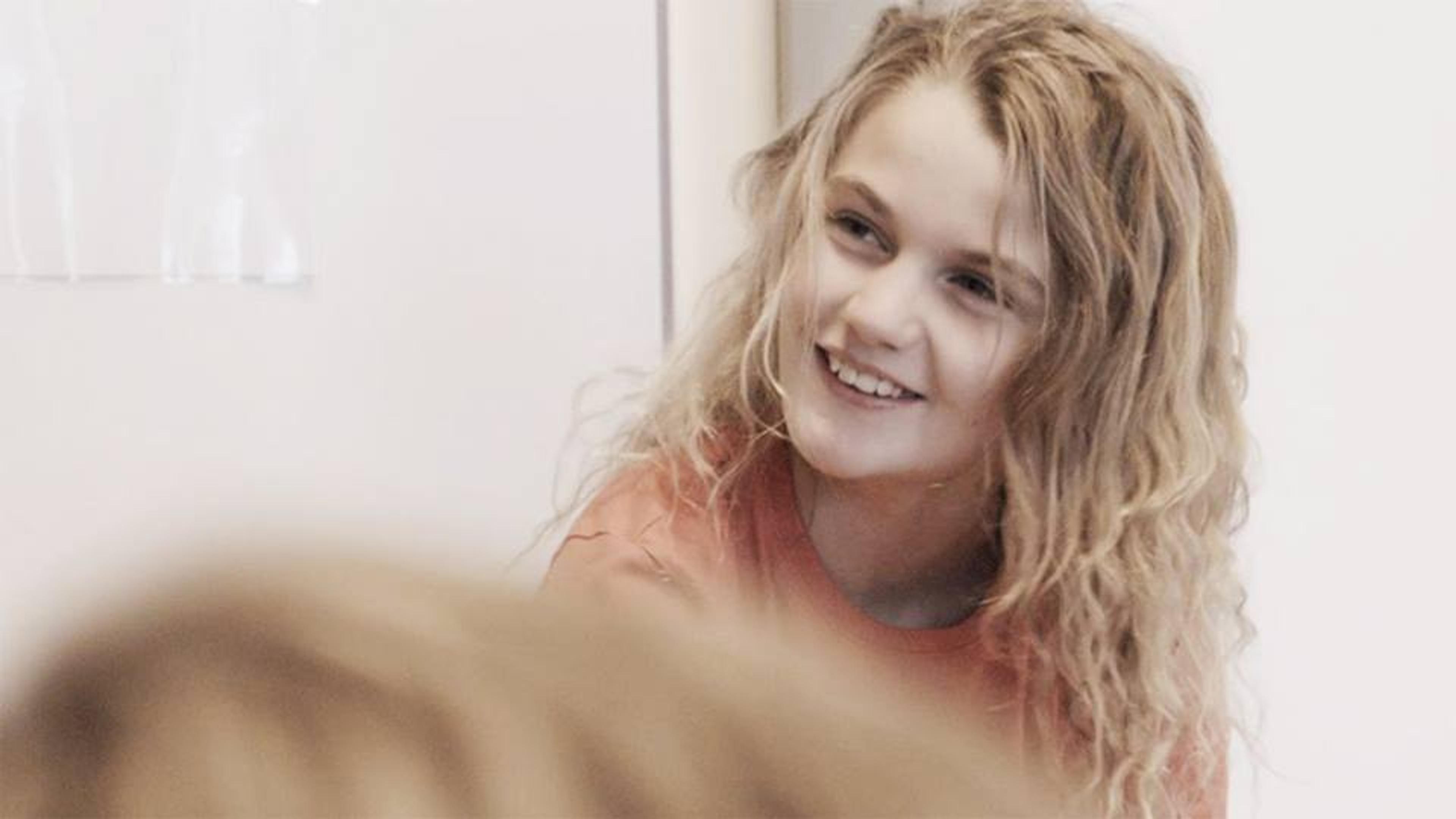 A smiling young person with long blonde wavy hair wearing a red shirt, looking slightly to the right.