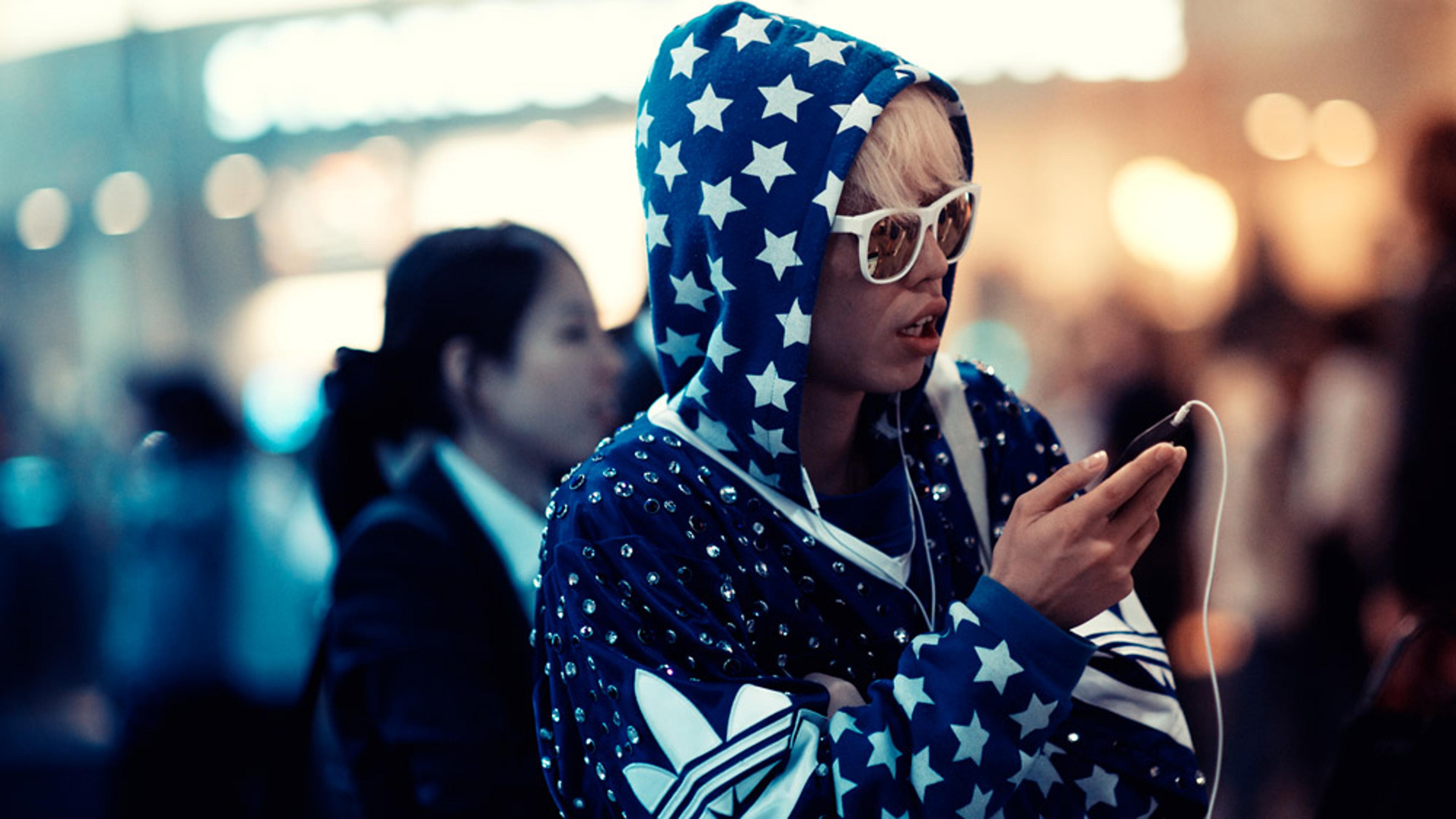 A woman in a navy starry hoodie and pink sunglasses using a phone with earbuds in a busy, softly blurred background.