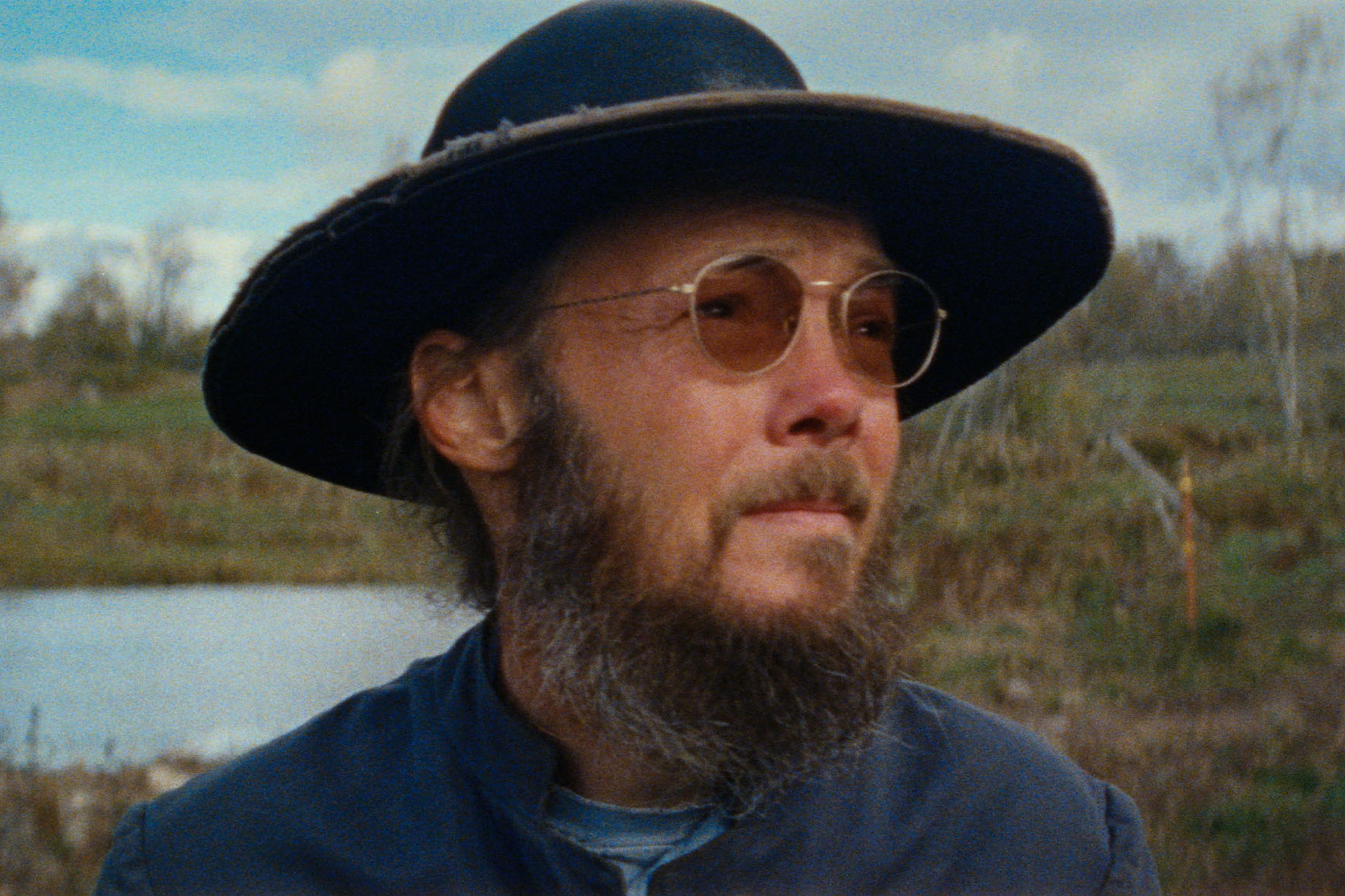 A man with a beard and glasses wearing a wide-brimmed hat and blue clothes stands outdoors near a pond, with trees in the background.