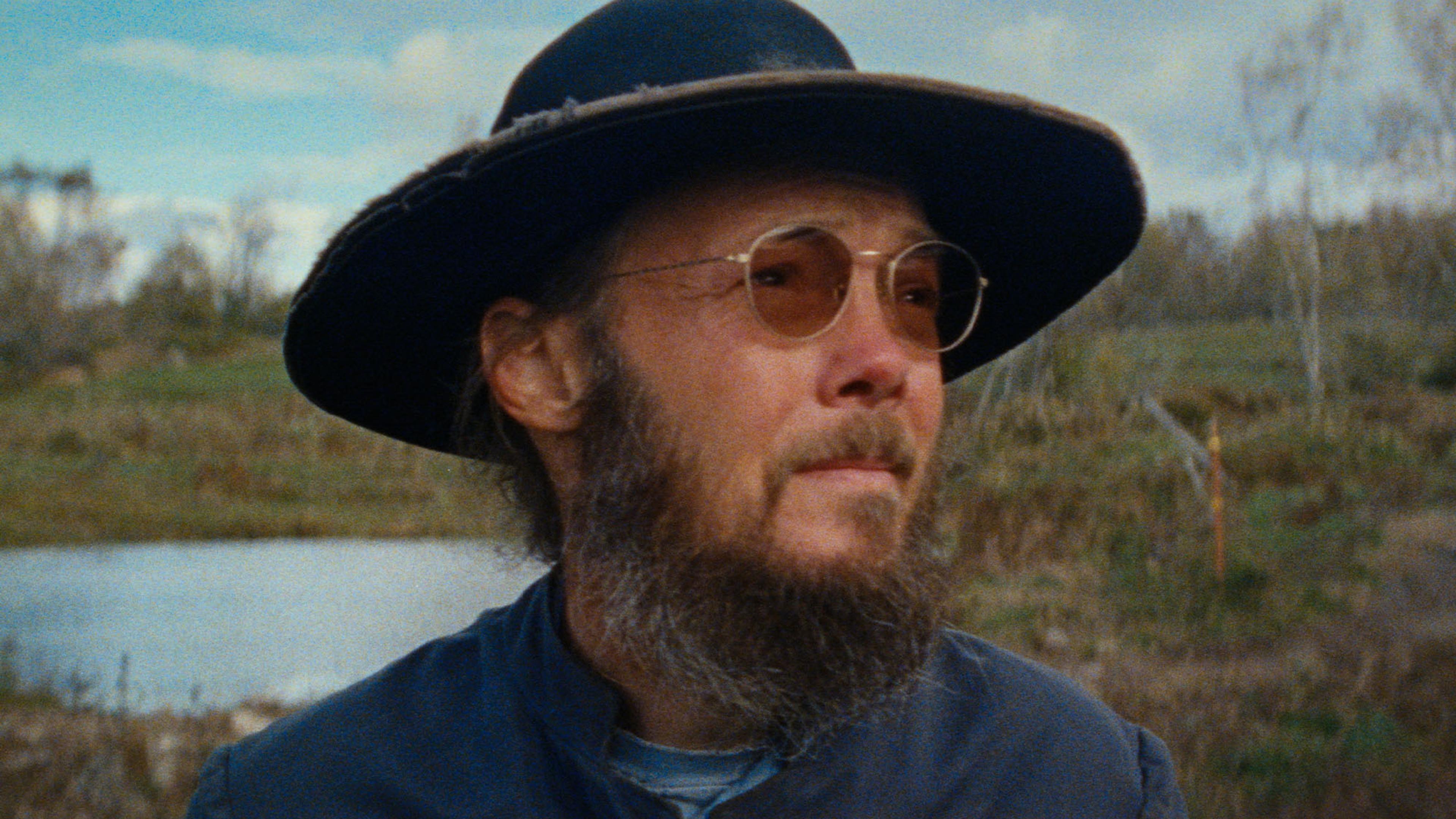 A man with a beard and glasses wearing a wide-brimmed hat and blue clothes stands outdoors near a pond, with trees in the background.