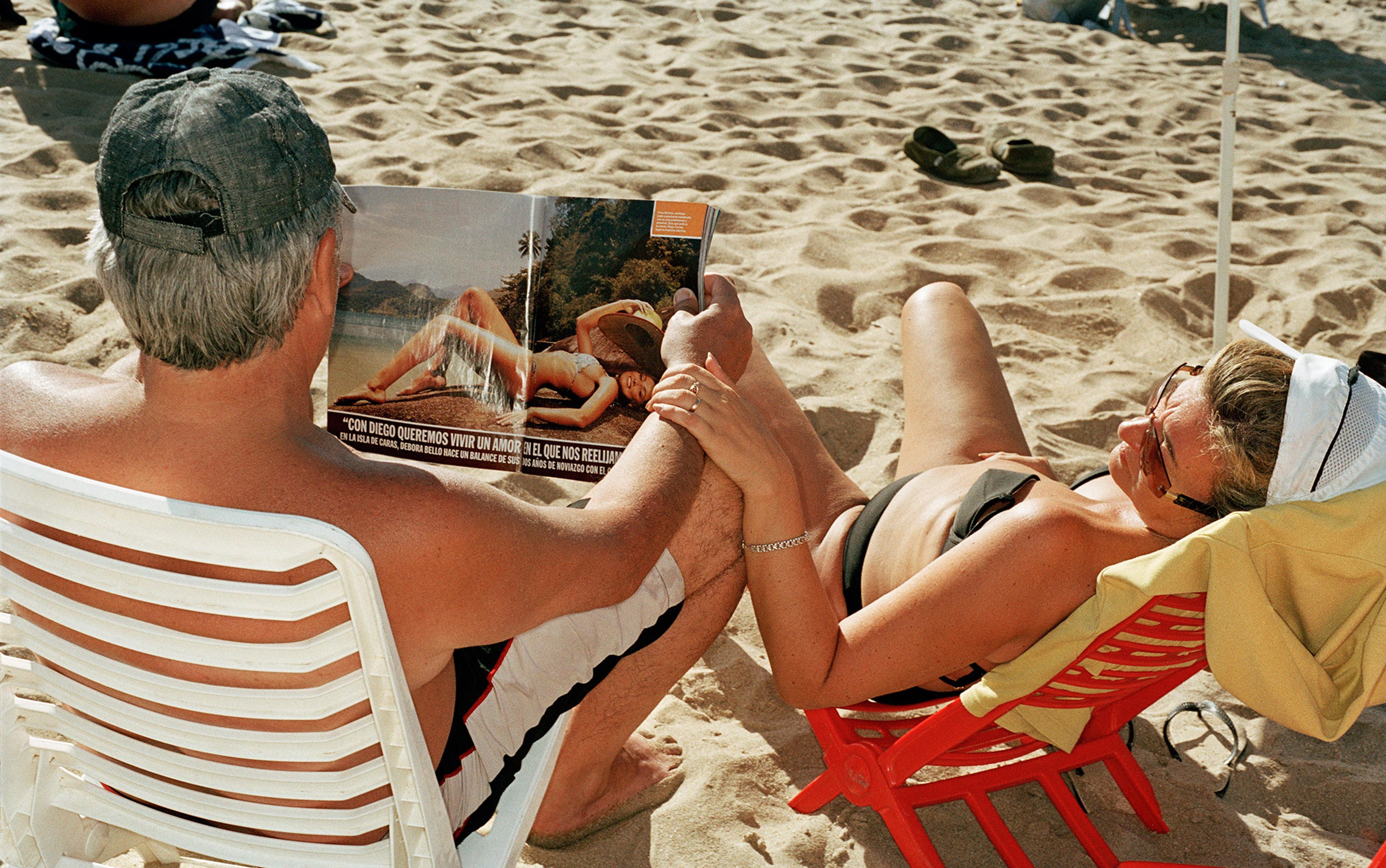 A man and a woman sunbathing on a beach, holding hands while the man reads a magazine open on a double-page spread of a reclining woman in a bikini.