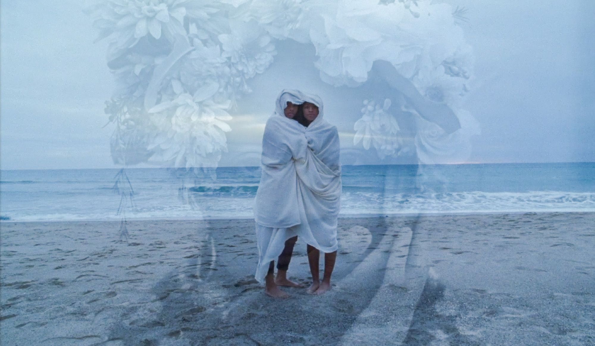 Two people wrapped in a blanket on a beach with a large floral overlay blending into the cloudy sky.