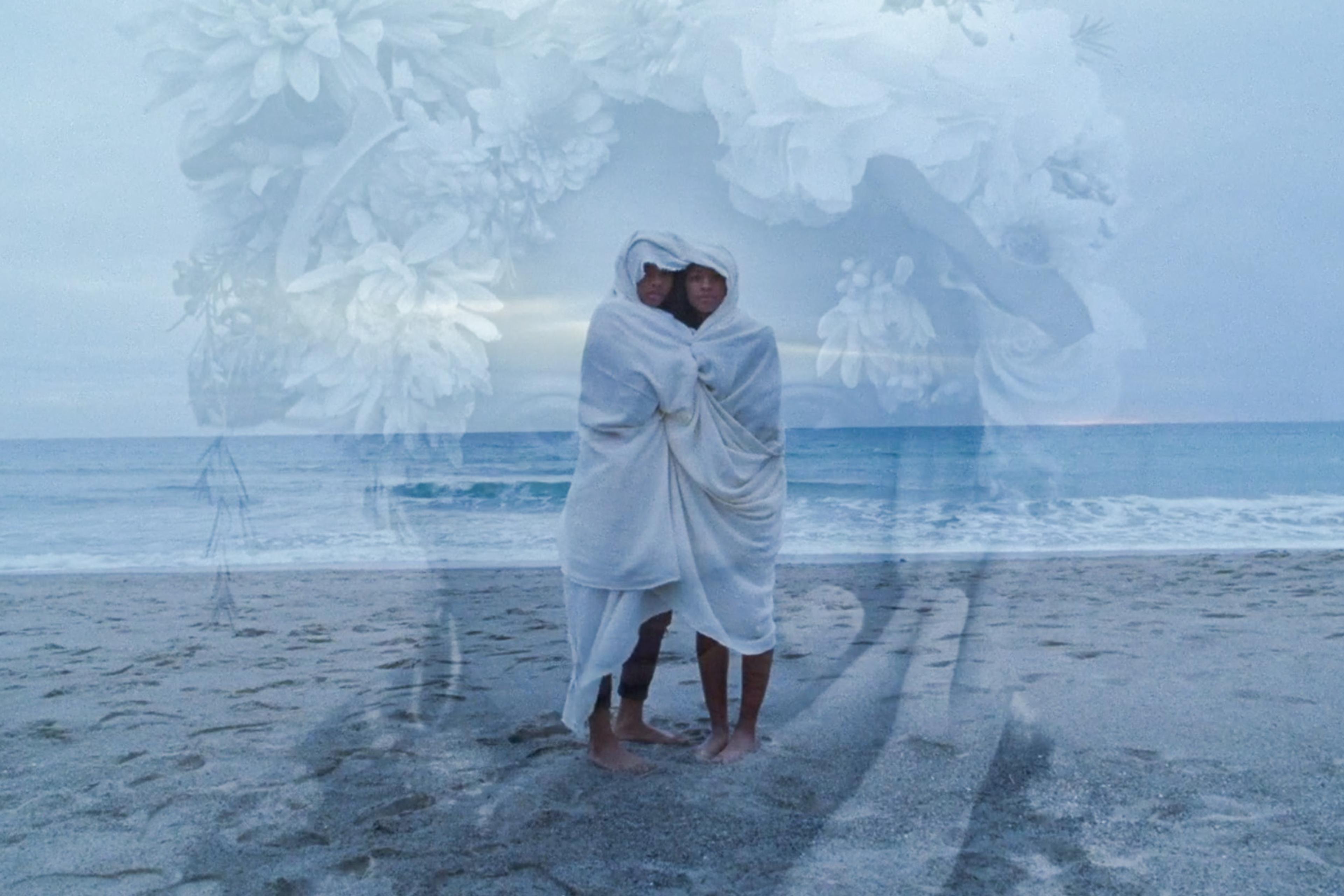 Two people wrapped in a blanket on a beach with a large floral overlay blending into the cloudy sky.