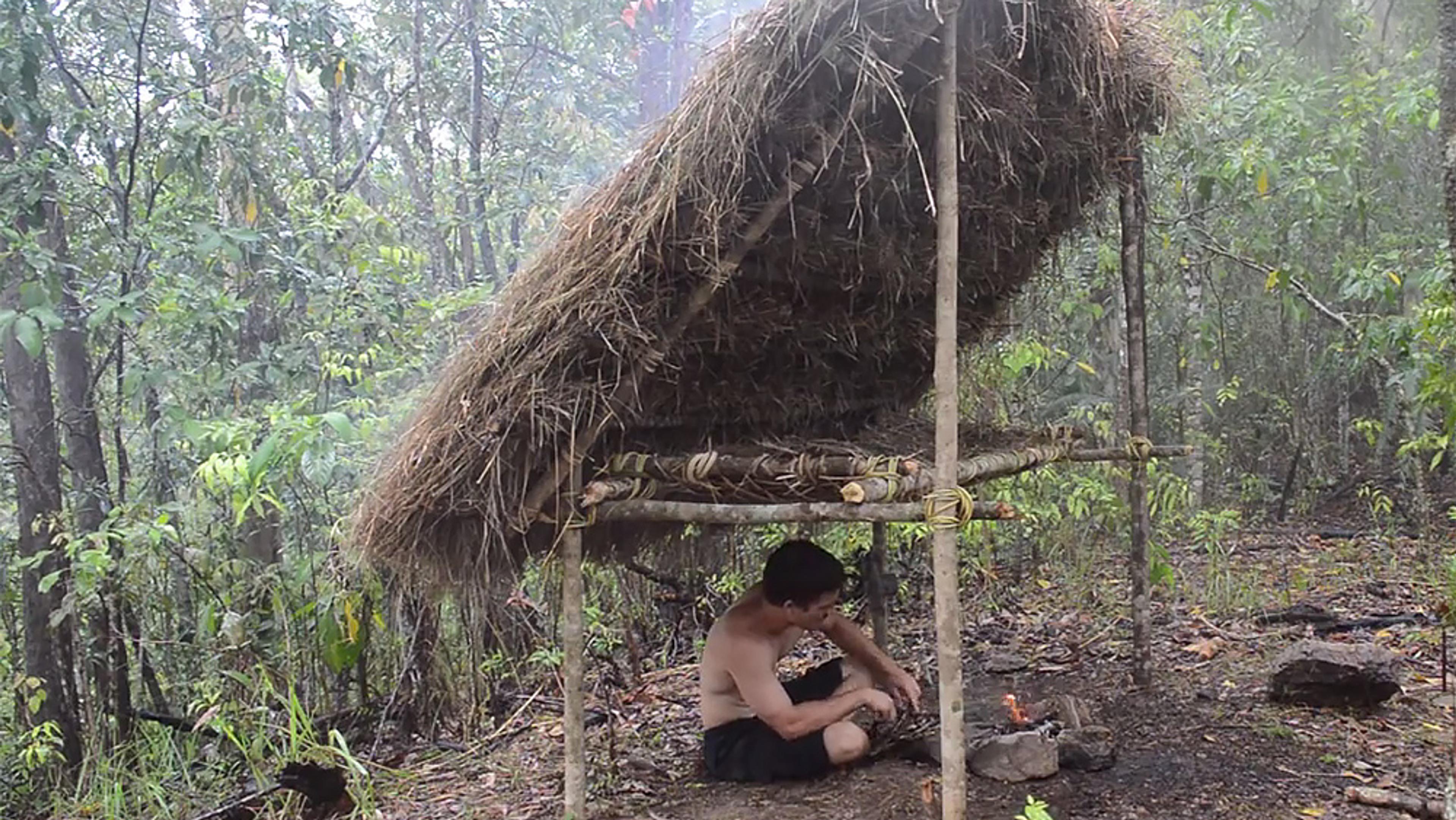 A shirtless man building a small fire under a shelter made of sticks and thatch in a dense forest. Smoke in the background.